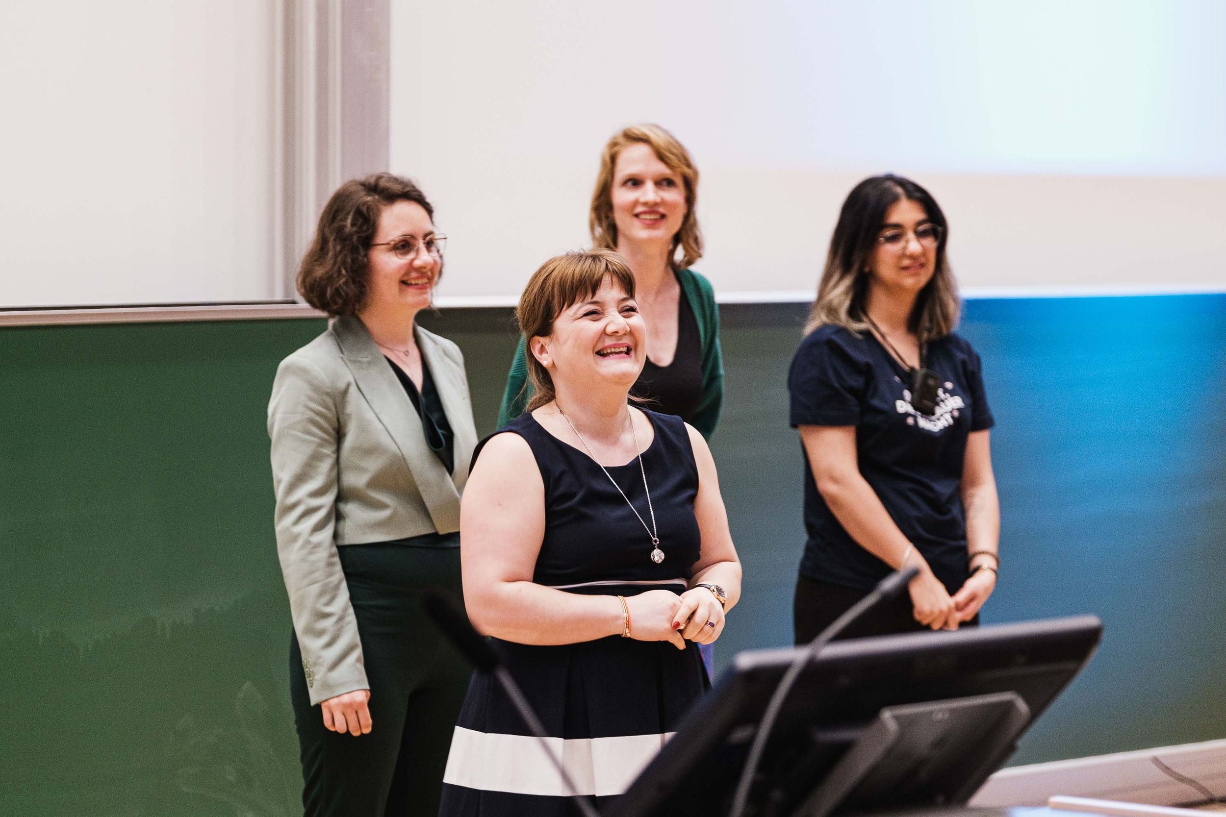Vier Frauen stehen im Klassenzimmer vor einer Tafel, lachen und sprechen. Eine Frau trägt ein schwarzes und weißes Kleid, die anderen tragen verschiedene blasse Kleidung. Im Vordergrund sieht man eine Computerkonsole.