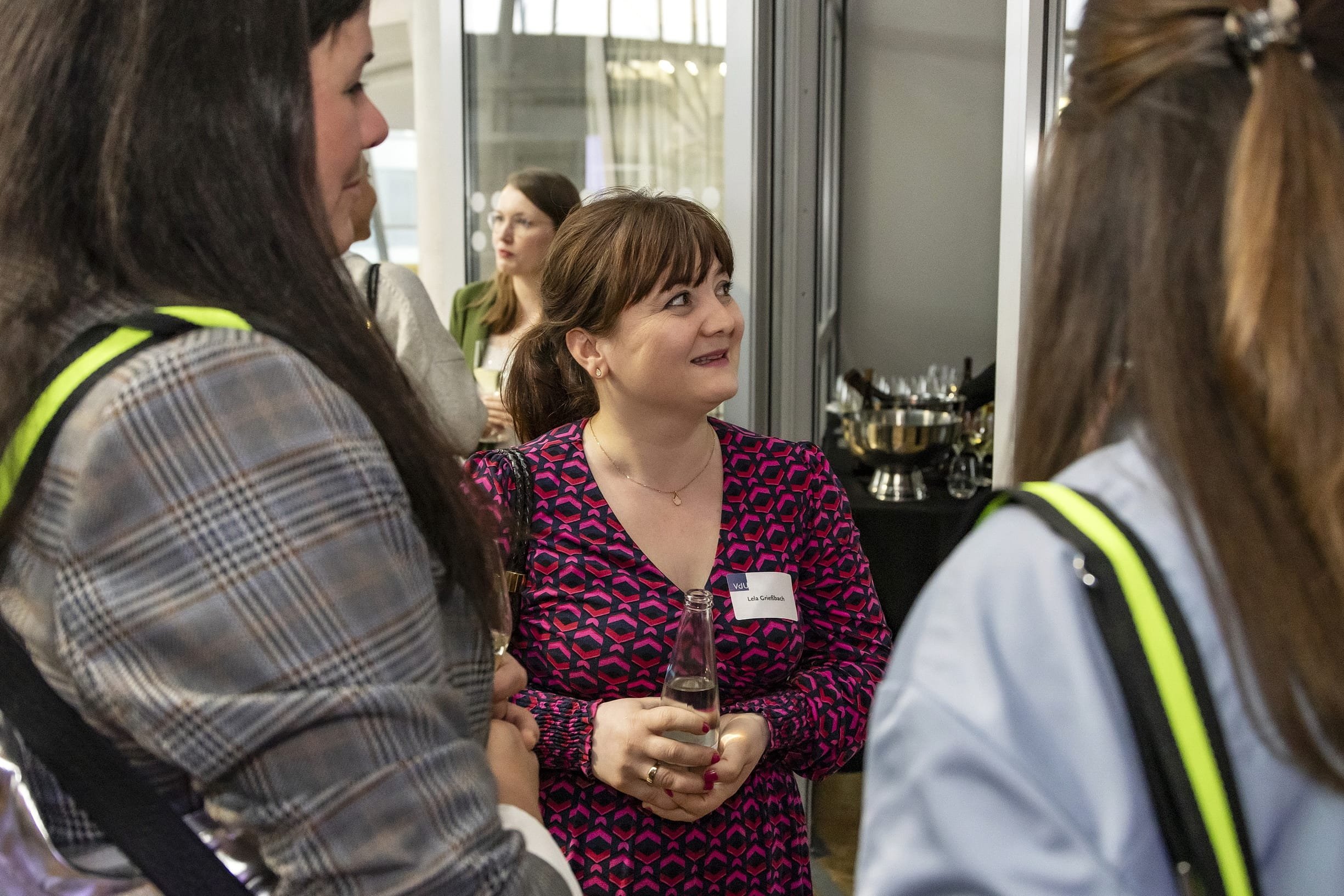 Mehrere Frauen im Gespräch bei einer Veranstaltung, eine Frau trägt ein pinkes, schwarzes Musterkleid und hält ein Getränk in der Hand.