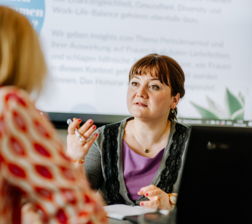 Frau spricht bei einer Präsentation vor einer Gruppe, im Hintergrund ein Bildschirm mit Text.