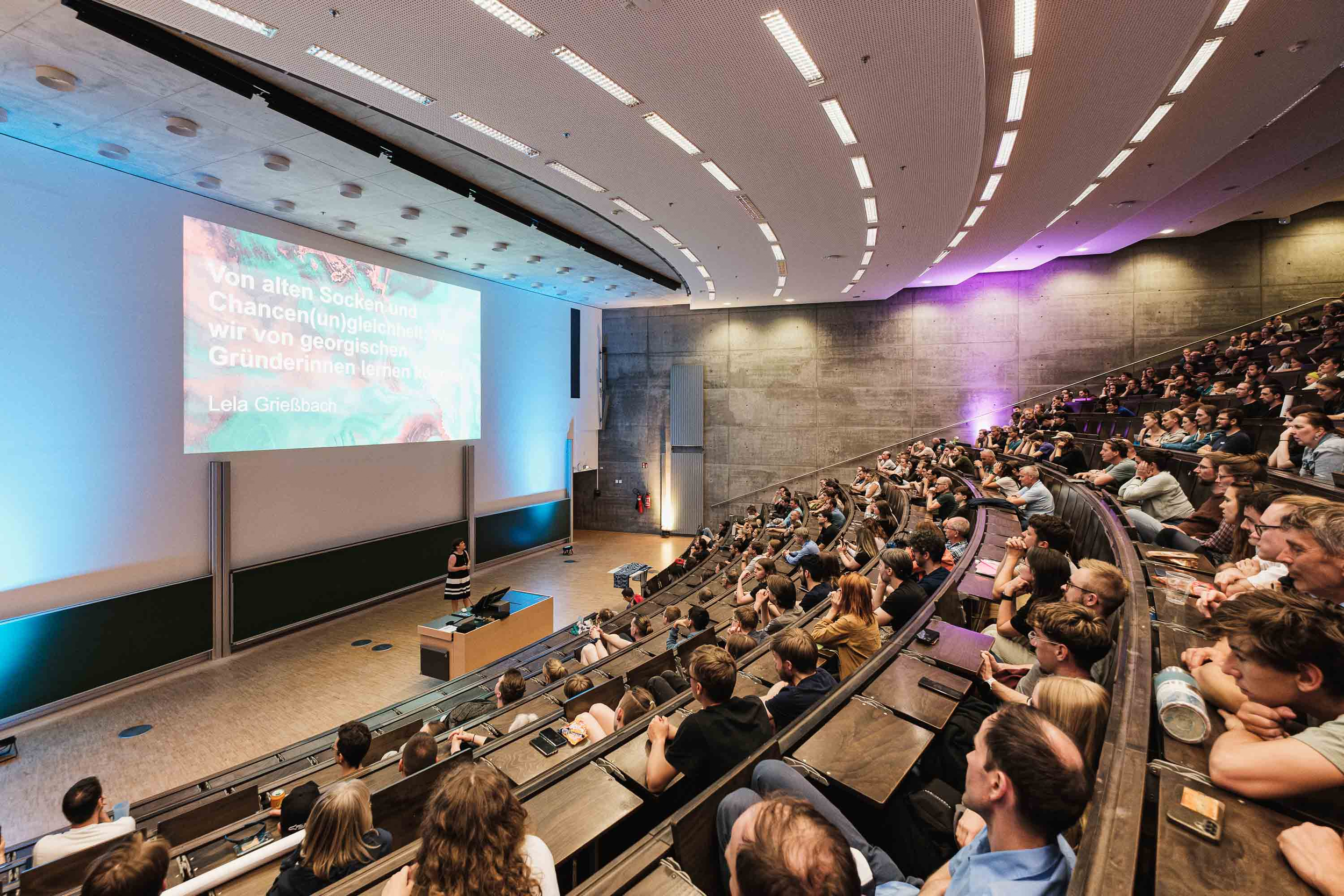 Ein volles Auditorium mit Menschen, die einer Präsentation in einem großen Hörsaal zuhören. Auf der Leinwand wird eine Folie mit Text auf Deutsch angezeigt.