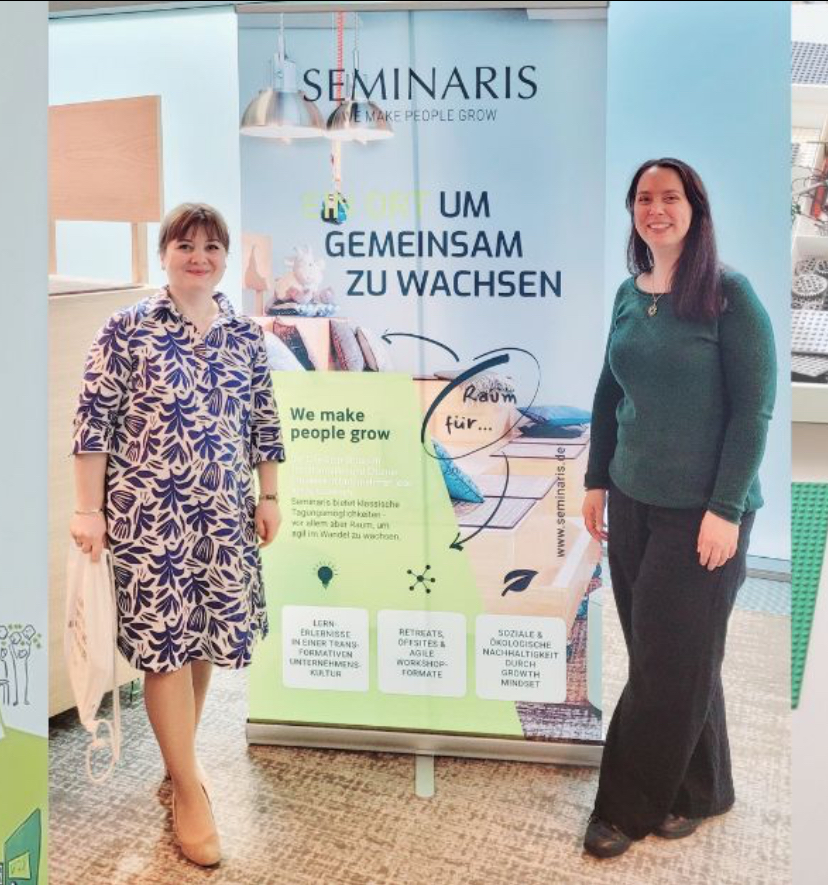 Zwei Frauen stehen neben einem Banner, das für ein Seminar wirbt, mit dem Thema 'Um gemeinsam zu wachsen'.