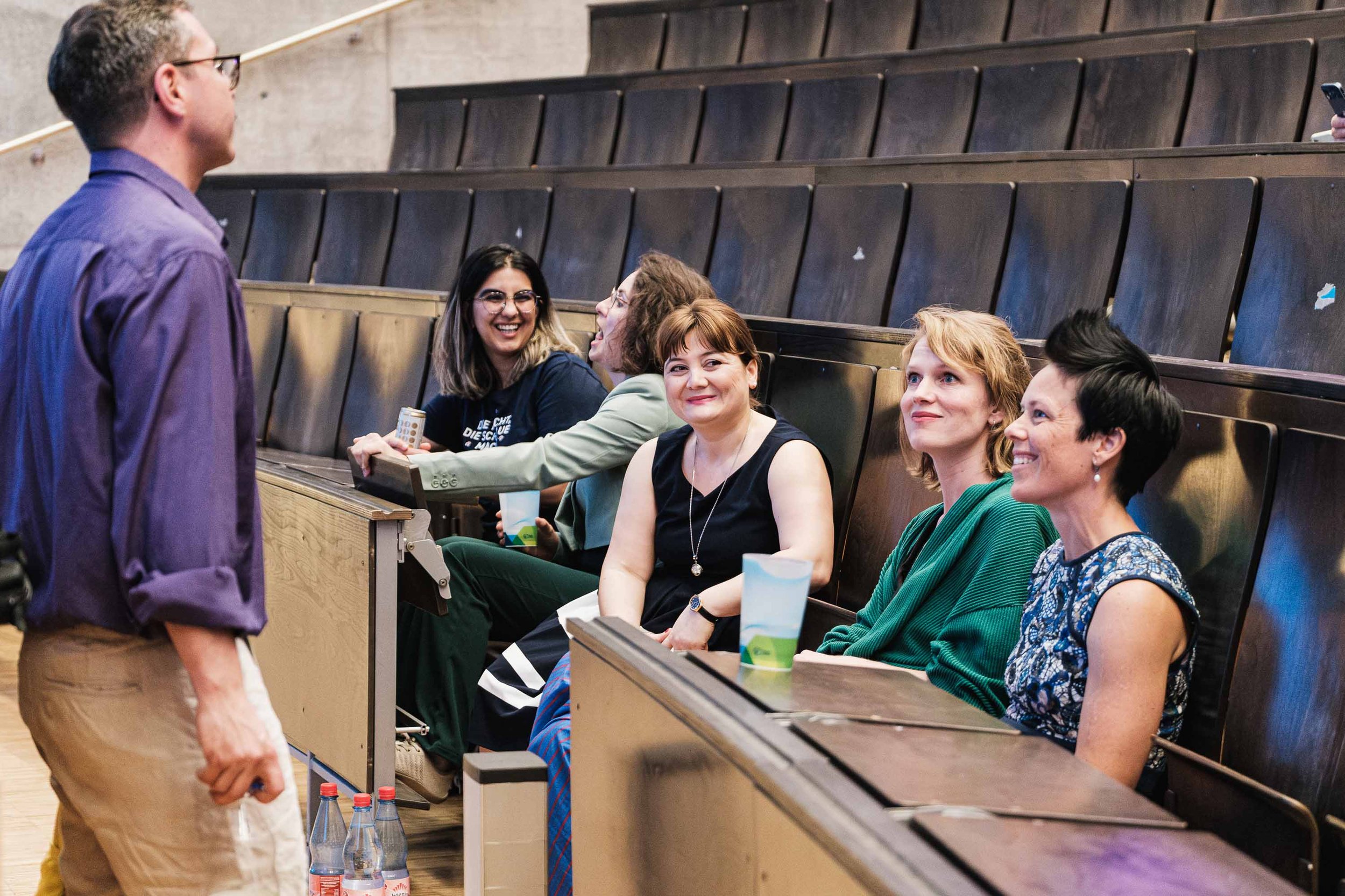 Fünf Frauen sitzen in einem Hörsaal und lachen, während ein Mann steht und spricht.