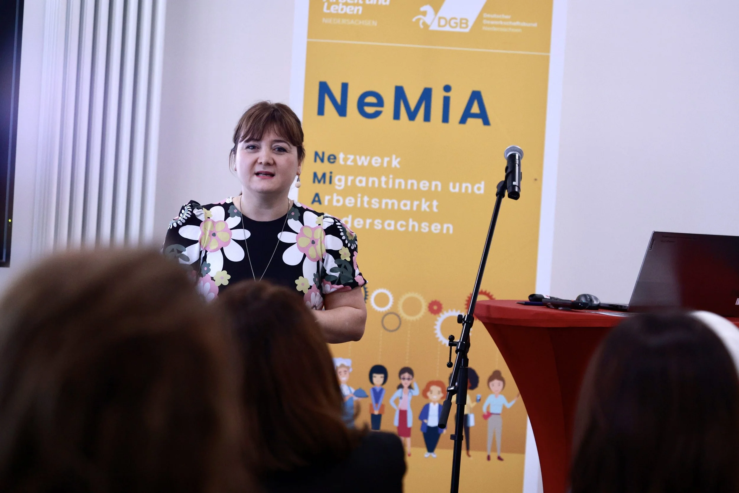 Eine Frau spricht auf einer Veranstaltung vor einem Publikum, mit einem gelben Banner im Hintergrund, das den Text 'NEMIA' und die Beschreibung 'Netzwerk Migrantinnen und Arbeitsmarkt Thüringen' zeigt.