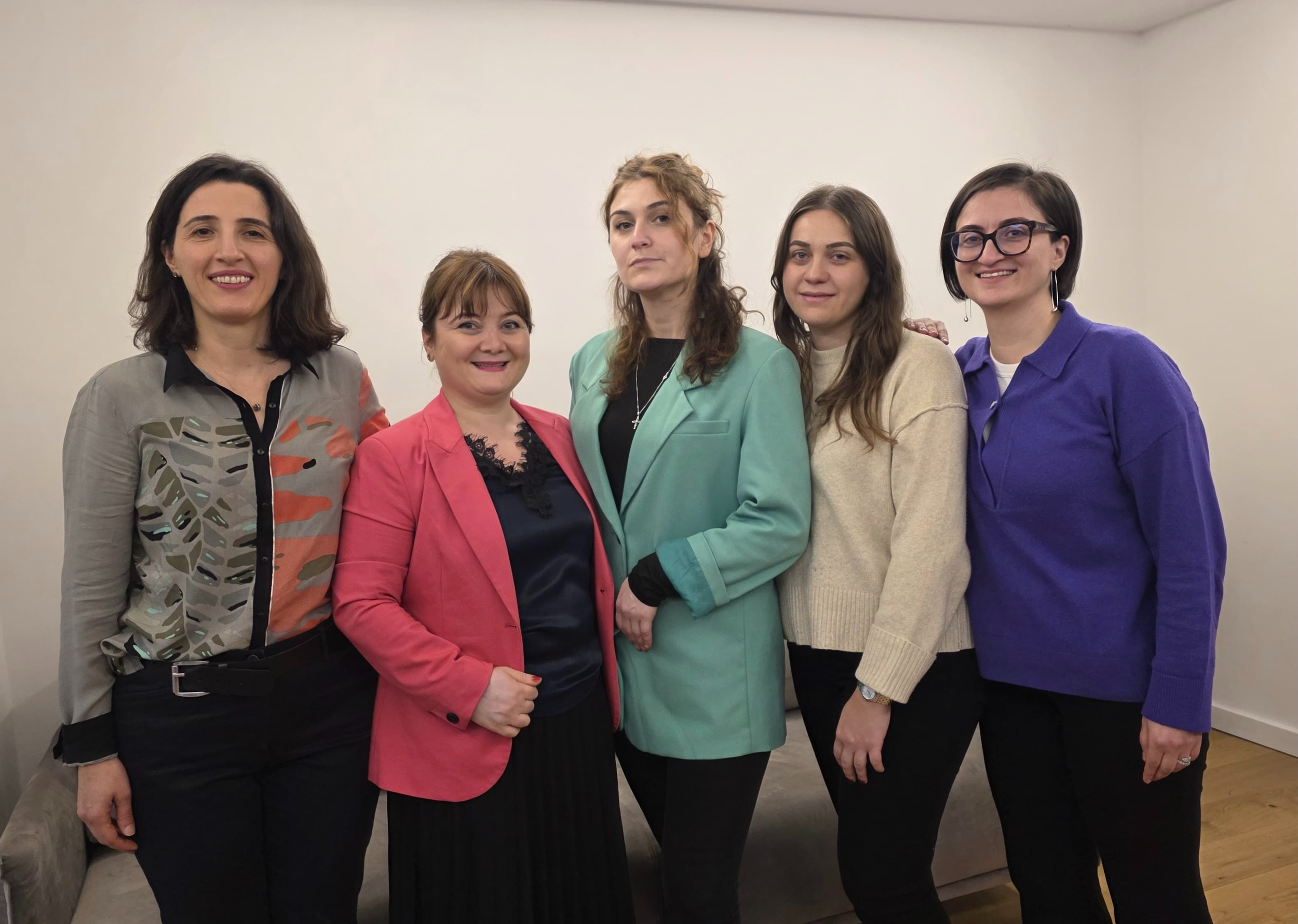 Fünf Frauen stehen zusammen in einem Raum, lächeln und tragen unterschiedliche bunte Jacken und Pullover.