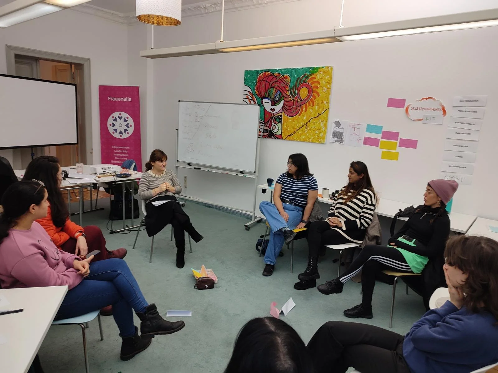 Mehrere Frauen sitzen in einem Kreis in einem Raum, der für eine Diskussion oder ein Seminar genutzt wird. Es gibt Pinnwände, einen Whiteboard mit Notizen und bunte Poster an den Wänden.