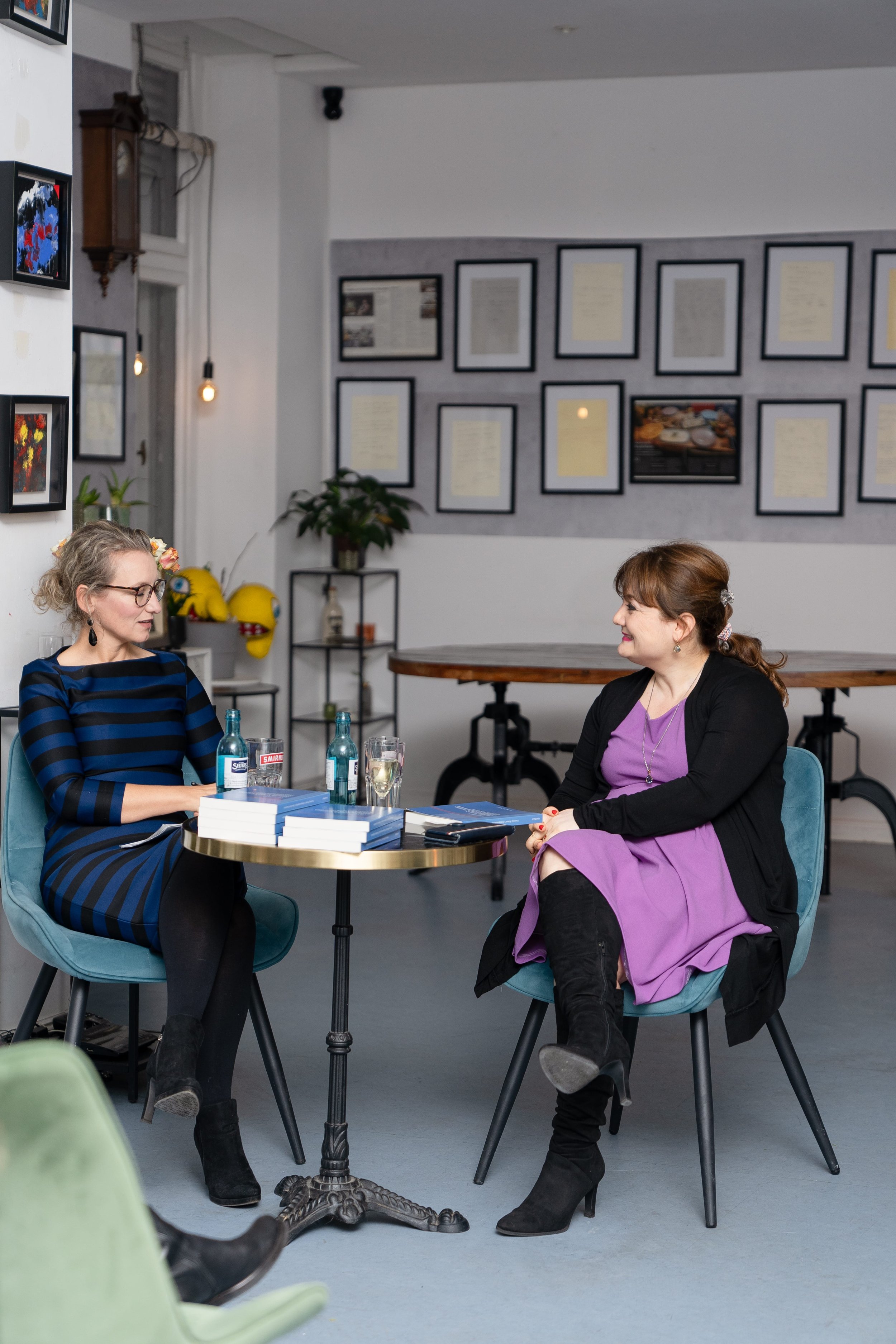 Zwei Frauen sitzen sich bei einem Gespräch in einem Café oder einer Buchhandlung gegenüber. Es liegen mehrere Bücher auf dem Tisch zwischen ihnen, zudem Wasser- und Glasflaschen. Im Hintergrund sind Bilder und Dokumente an der Wand, eine Pflanze und dekorative Gegenstände sichtbar.