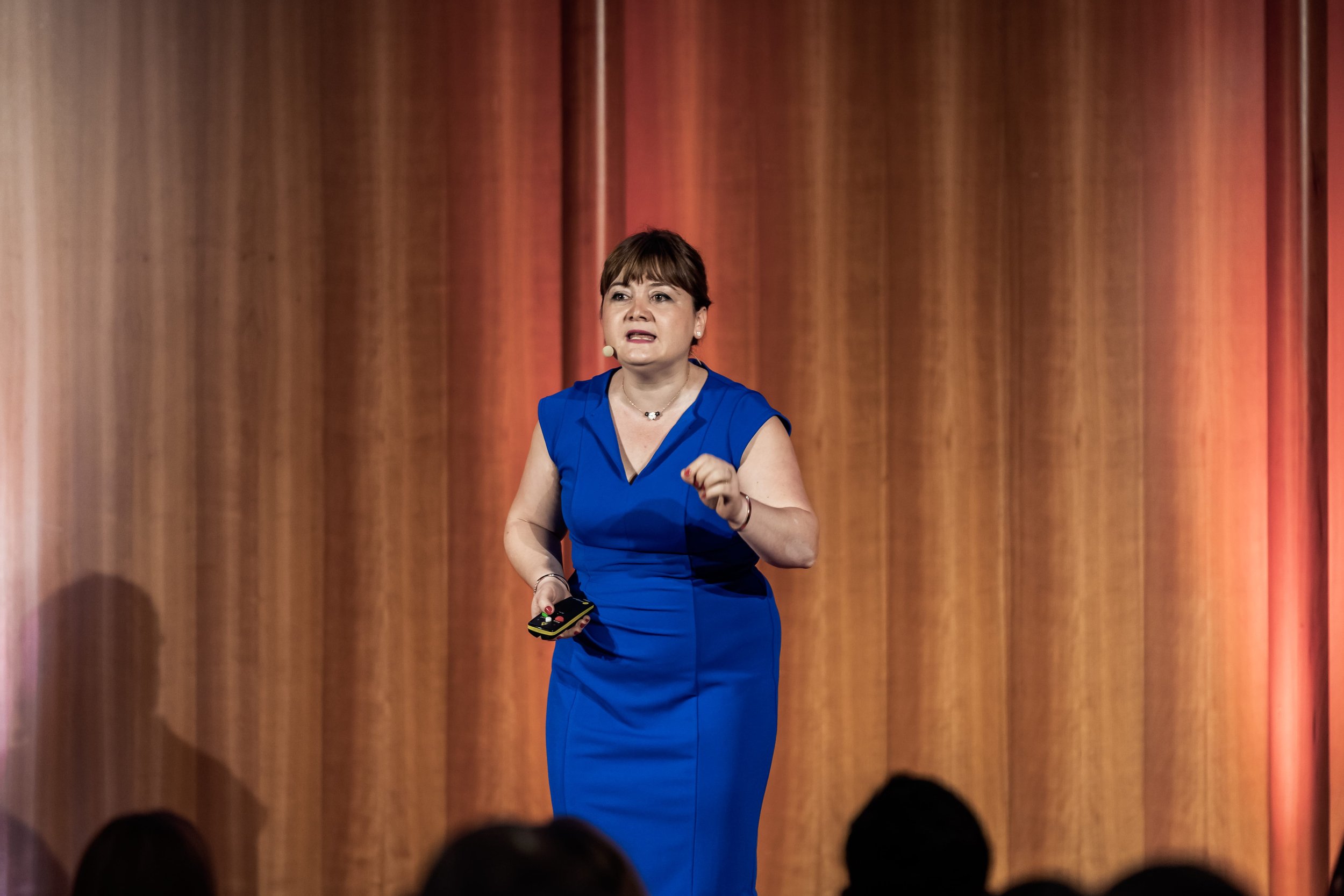 Eine Frau in einem blauen Kleid, die auf einer Bühne vor einem orangfarbenen Vorhang spricht, mit einem Mikrofon im Ohr und einem Handgerät in der Hand.