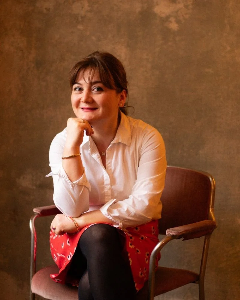 Eine Frau sitzt auf einem Stuhl, trägt eine weiße Bluse und einen roten Rock mit schwarzer Strumpfhose, hat braune Haare und lächelt.