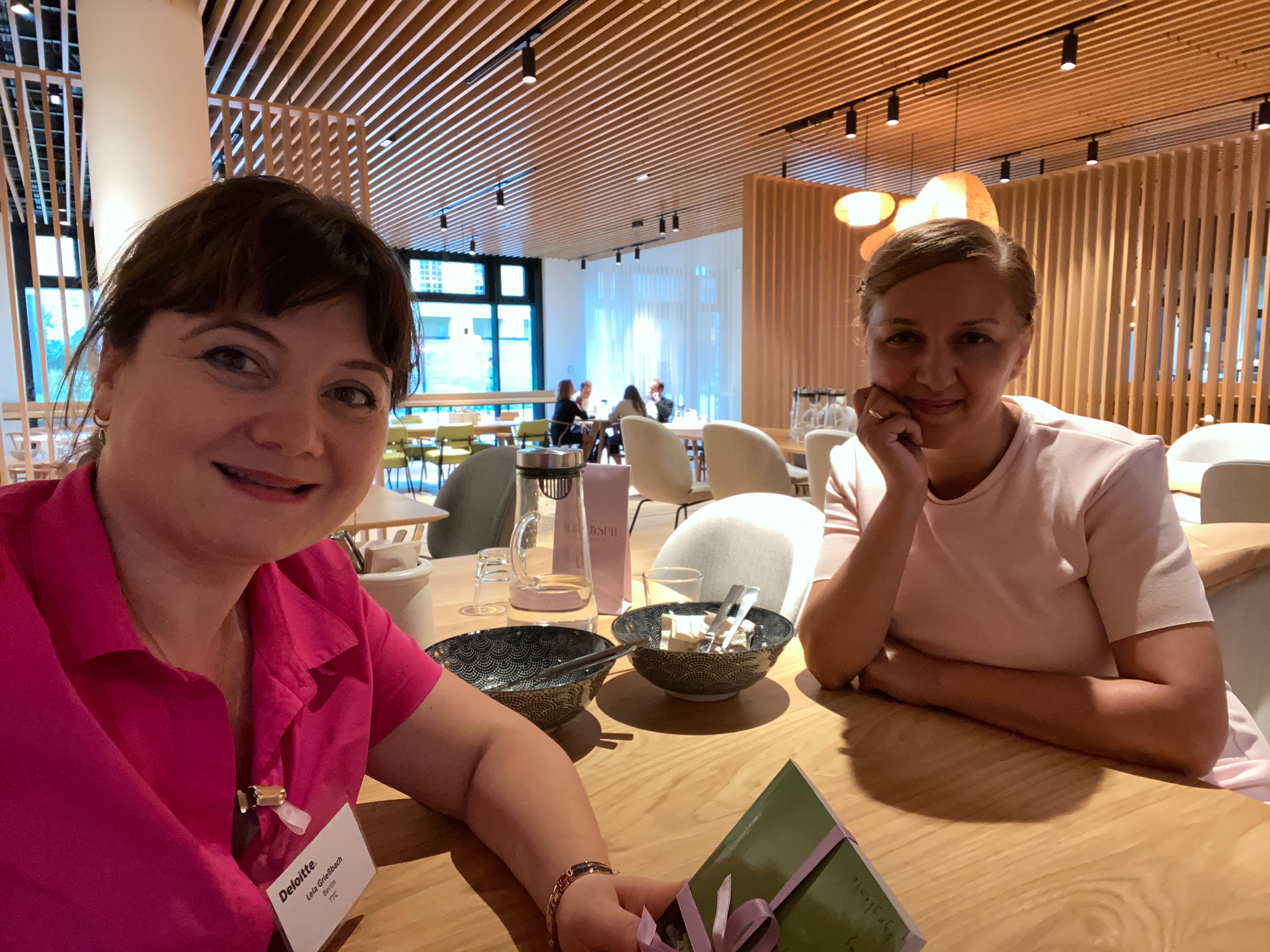 Zwei Frauen sitzen an einem Tisch in einem modernen Restaurant, lächeln. Im Hintergrund sitzen weitere Gäste, das Restaurant hat Holzdecke und wechselnde Beleuchtung.