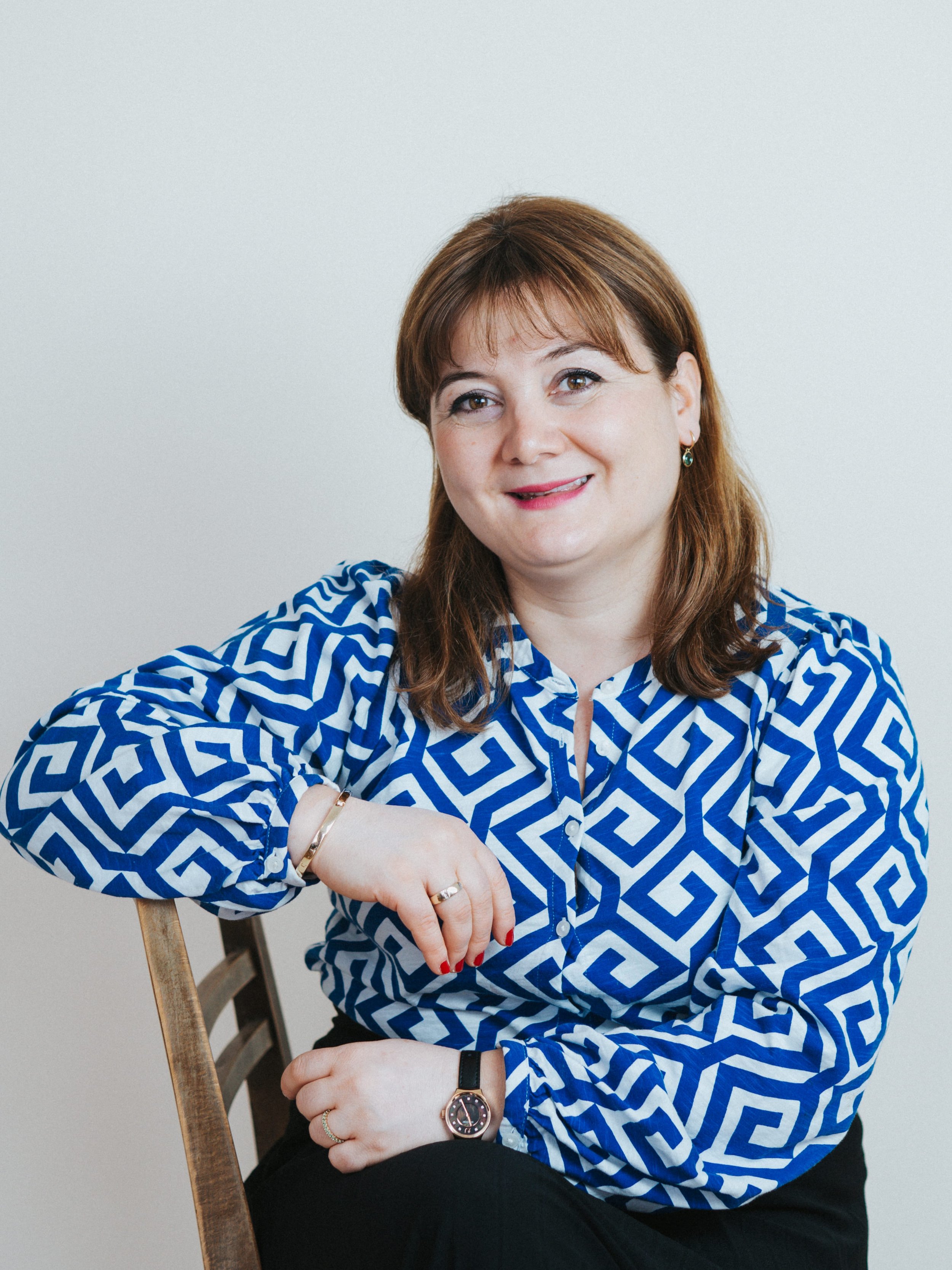 Eine Frau mit braunen Haaren, die ein blauen und weißen geometrischen Muster Hemd trägt, sitzt auf einem Stuhl gegen einen weißen Hintergrund.