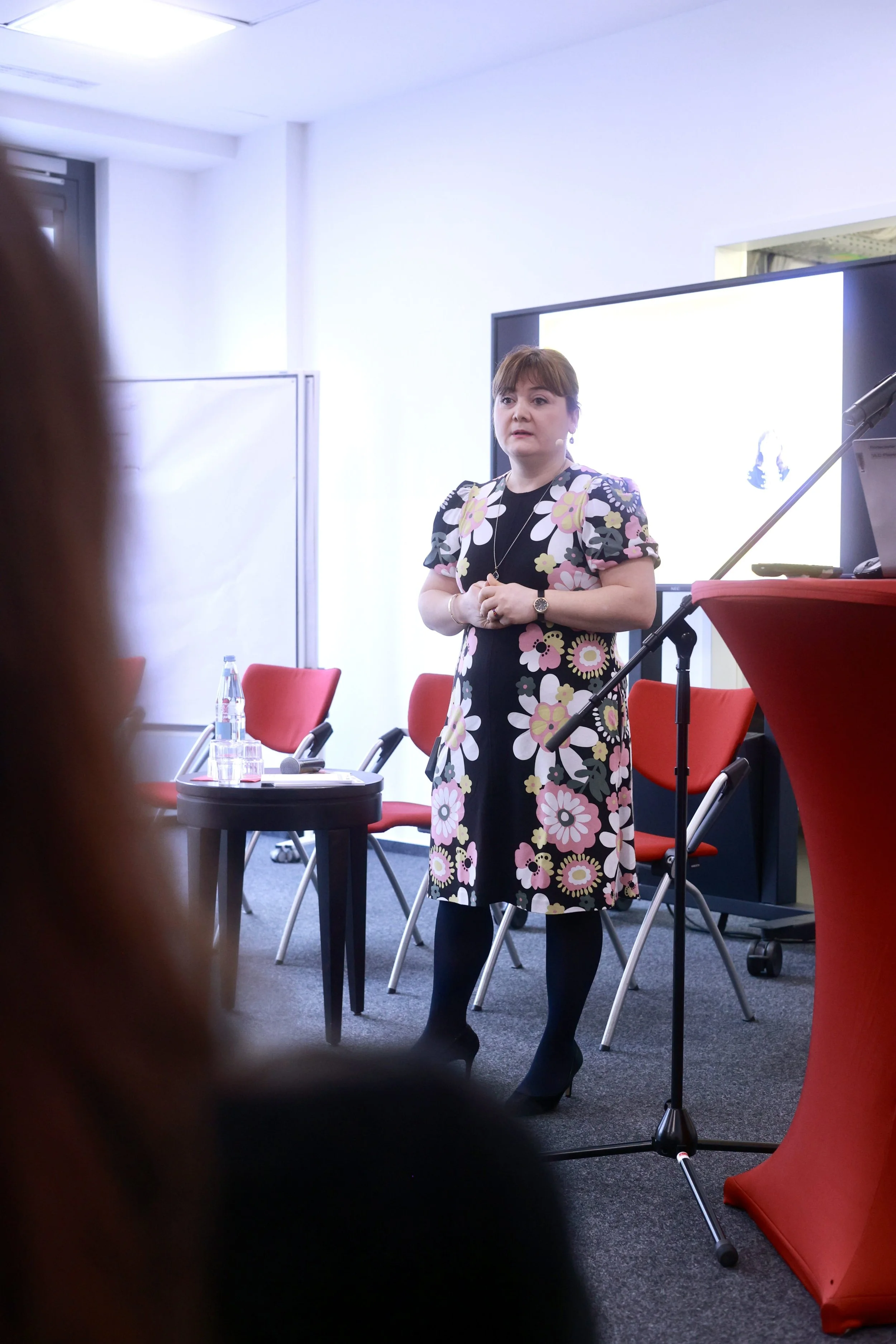 Eine Frau steht vor einem Publikum, spricht bei einer Veranstaltung, im Hintergrund ein Bildschirm, rote Stühle, Wasserflaschen auf einem Tisch