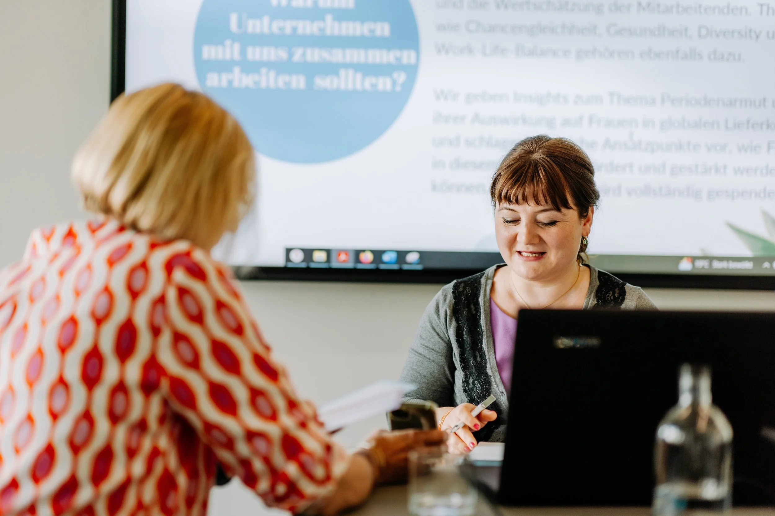 Zwei Frauen in einer Präsentation, eine blickt auf ihr Smartphone, die andere lächelt. Im Hintergrund ein großer Bildschirm mit einem blauen Titelblock und Text in deutscher Sprache.