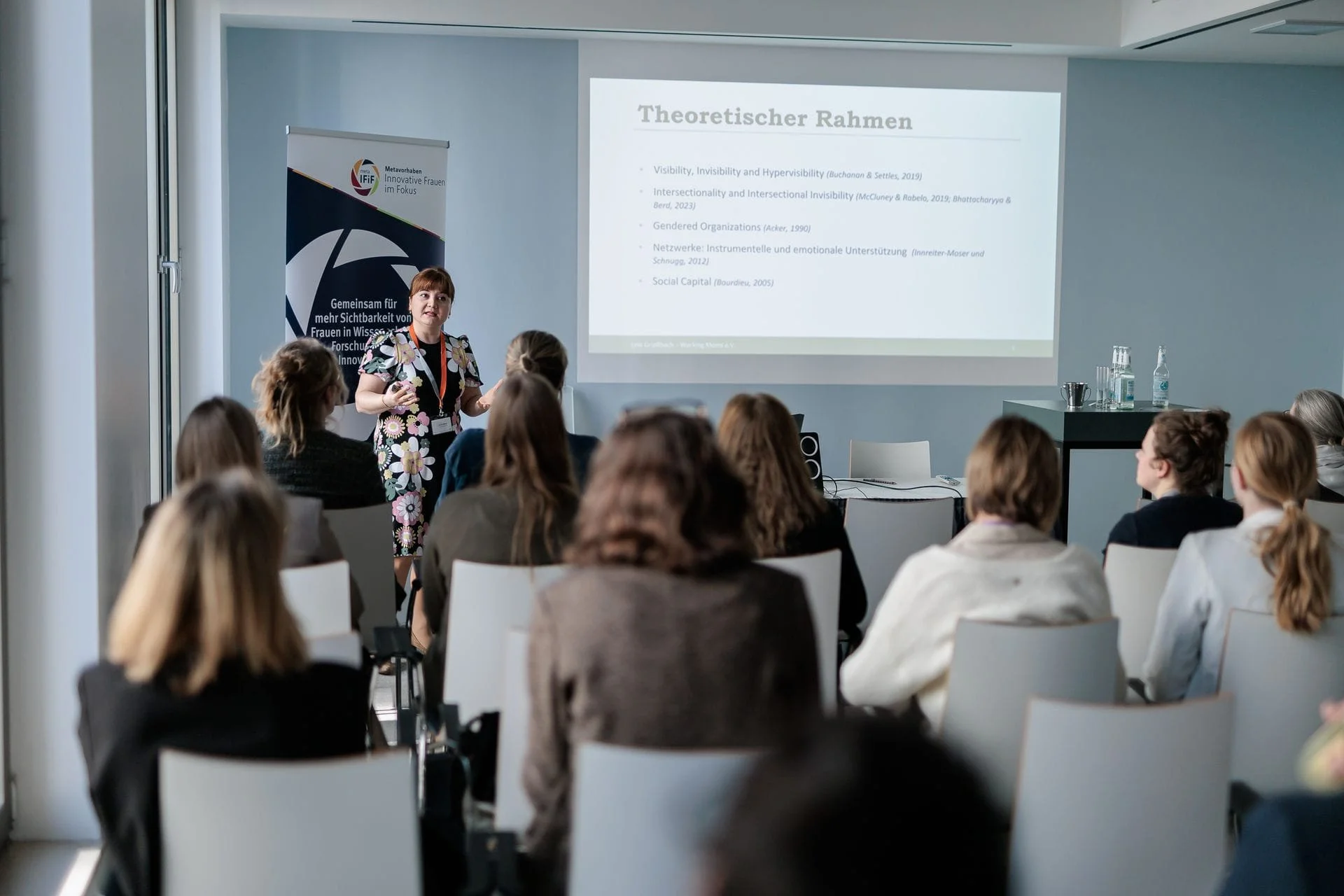 Eine Frau hält eine Präsentation vor einem Publikum in einem modernen Konferenzraum. Im Hintergrund ist eine Projektionsfolie mit dem Titel 'Theoretischer Rahmen' und einigen Referenzen zu sehen. Das Publikum besteht aus Frauen, die aufmerksam zuhören.