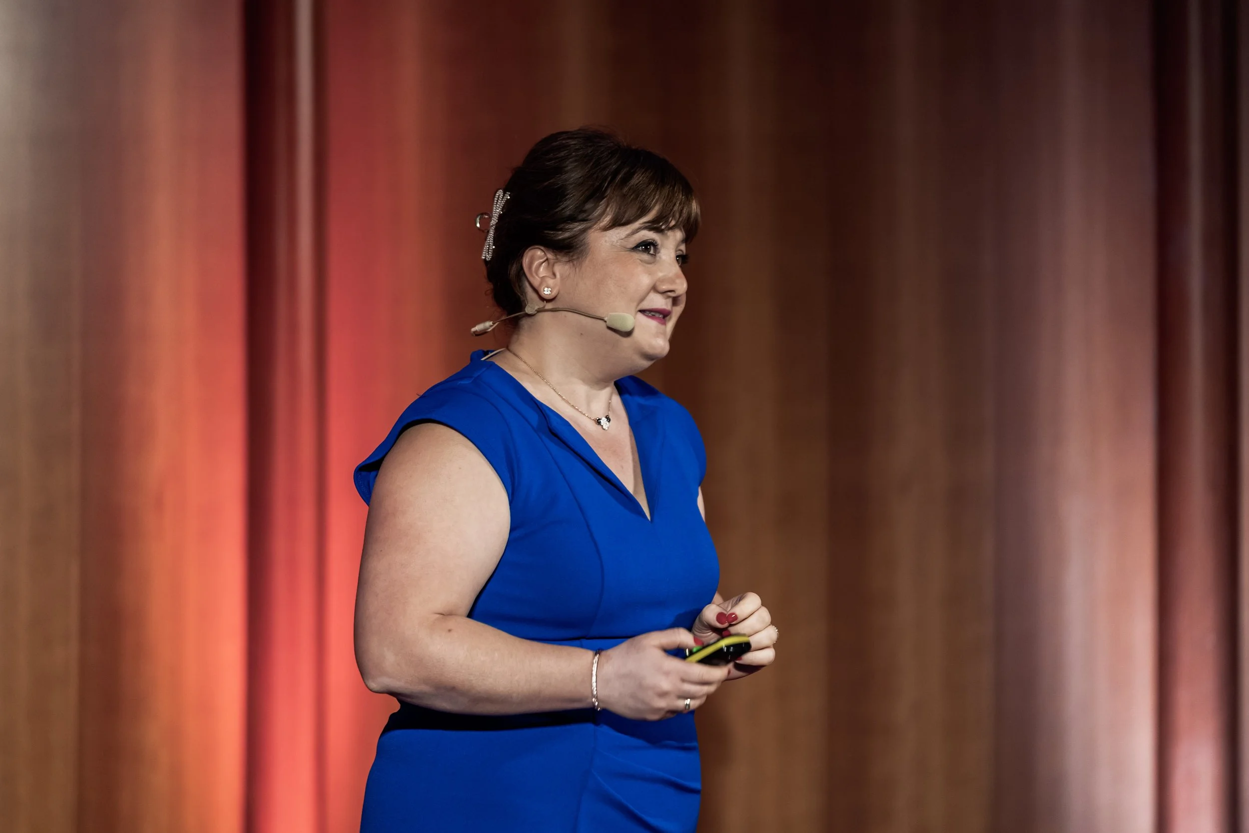 Frau spricht bei einem Vortrag, trägt ein blaues Kleid, hält ein Smartphone, trägt einen Headset, vor einem roten Vorhang.
