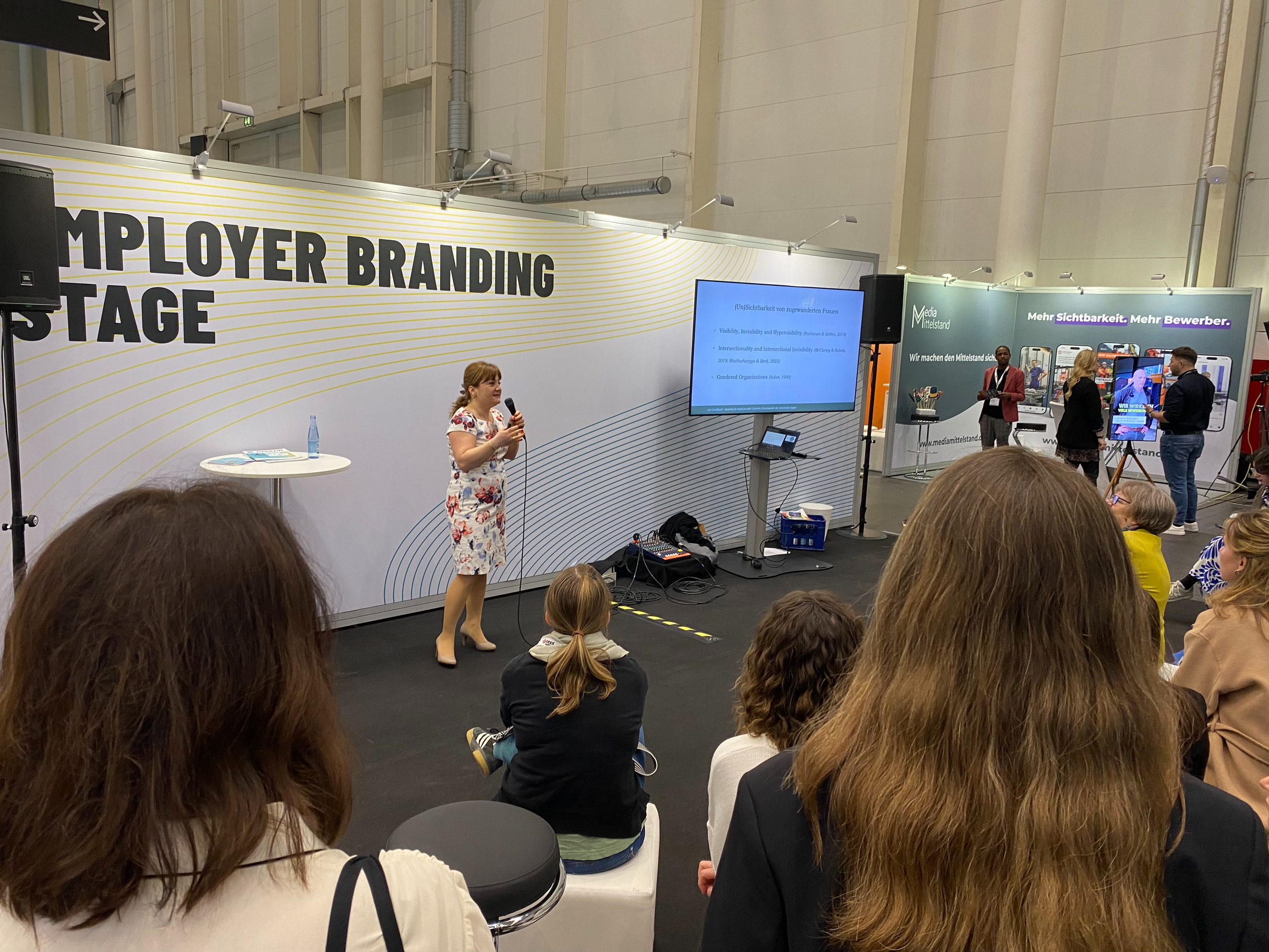 Frau spricht auf einer Bühne bei einer Veranstaltung namens 'Employer Branding Tage'. Es sitzen mehrere Menschen, die ihr zuhören. Im Hintergrund sind Beschilderungen und Bildschirme.