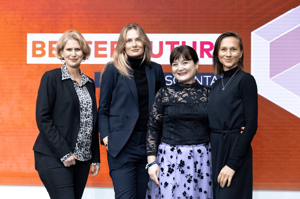 Vier Frauen stehen vor einem orangefarbenen Hintergrund mit Text, die lächeln.