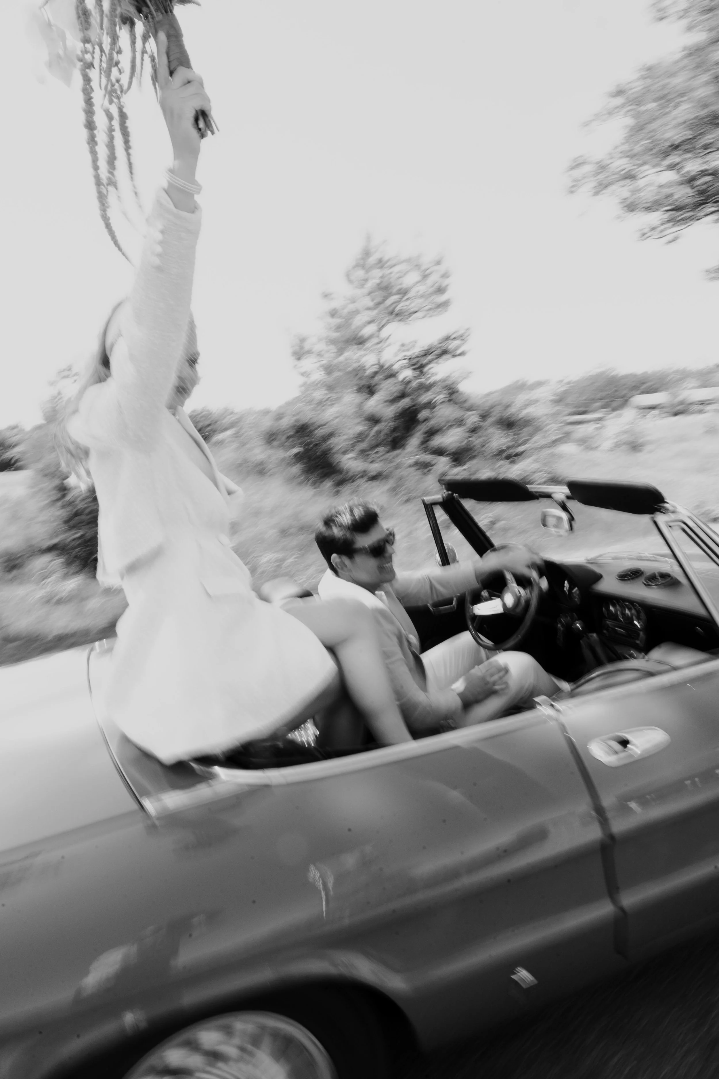 Een paar in een open auto rijden door een landschap met bomen, de vrouw zit op de rand van de auto met haar benen buiten de auto, terwijl de man achter het stuur zit.