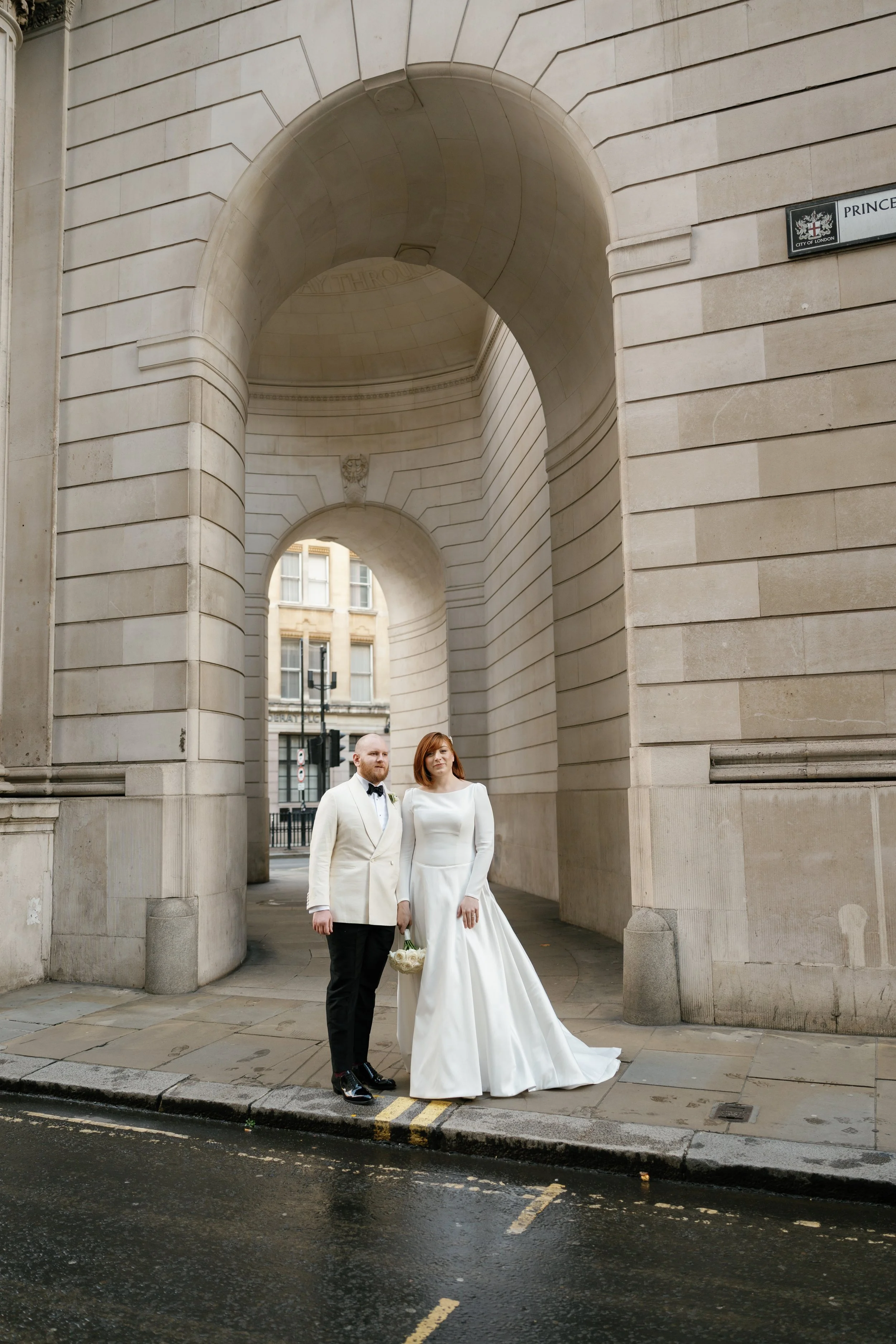 Een pasgetrouwd paar in formele trouwkleding poseert onder een grote boog in Londen, met een stadsmuur op de achtergrond.