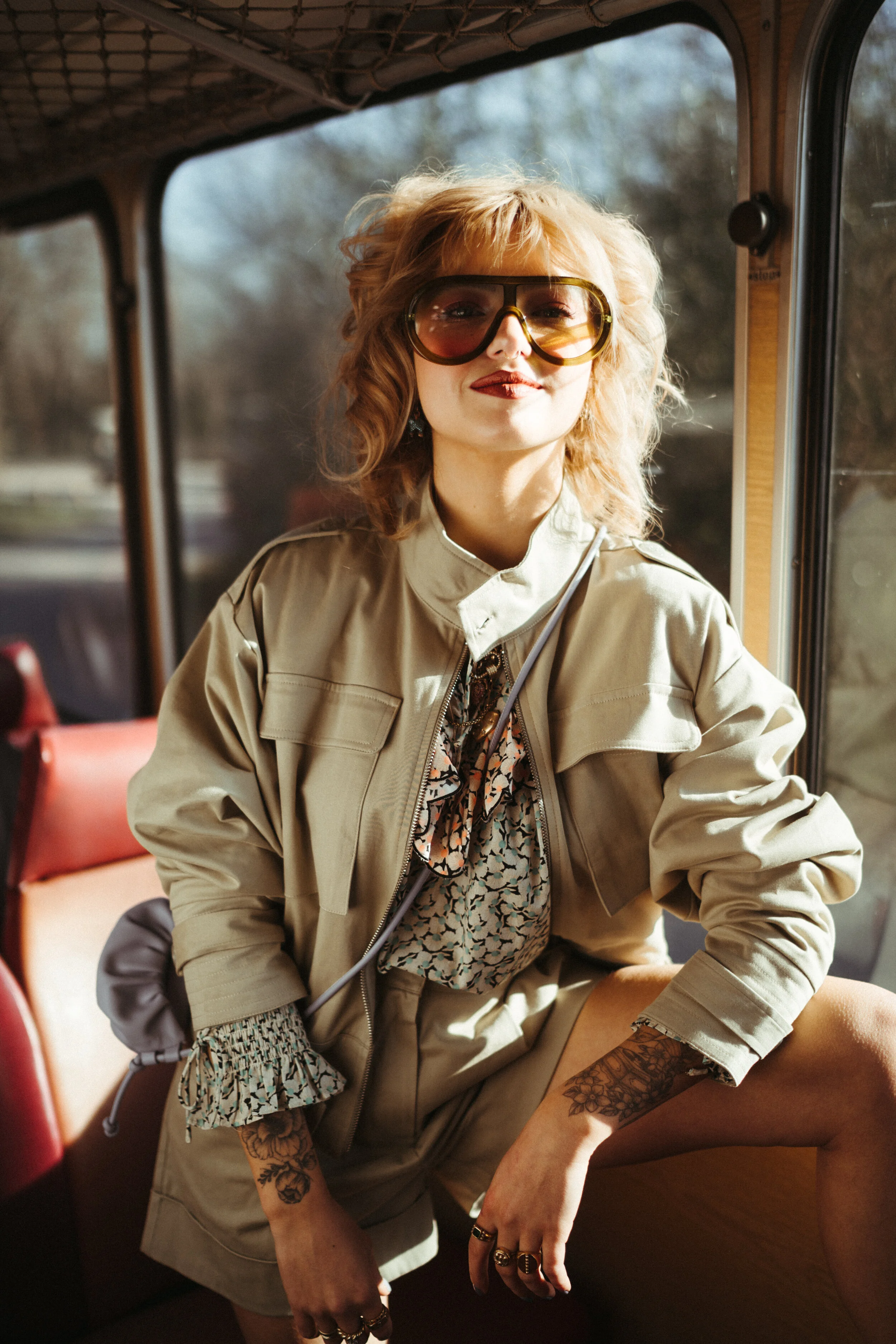 Een vrouw met rood, golvend haar draagt grote, gele zonnebril en een beige jas, poseert in een bus met uitzicht op de achtergrond.