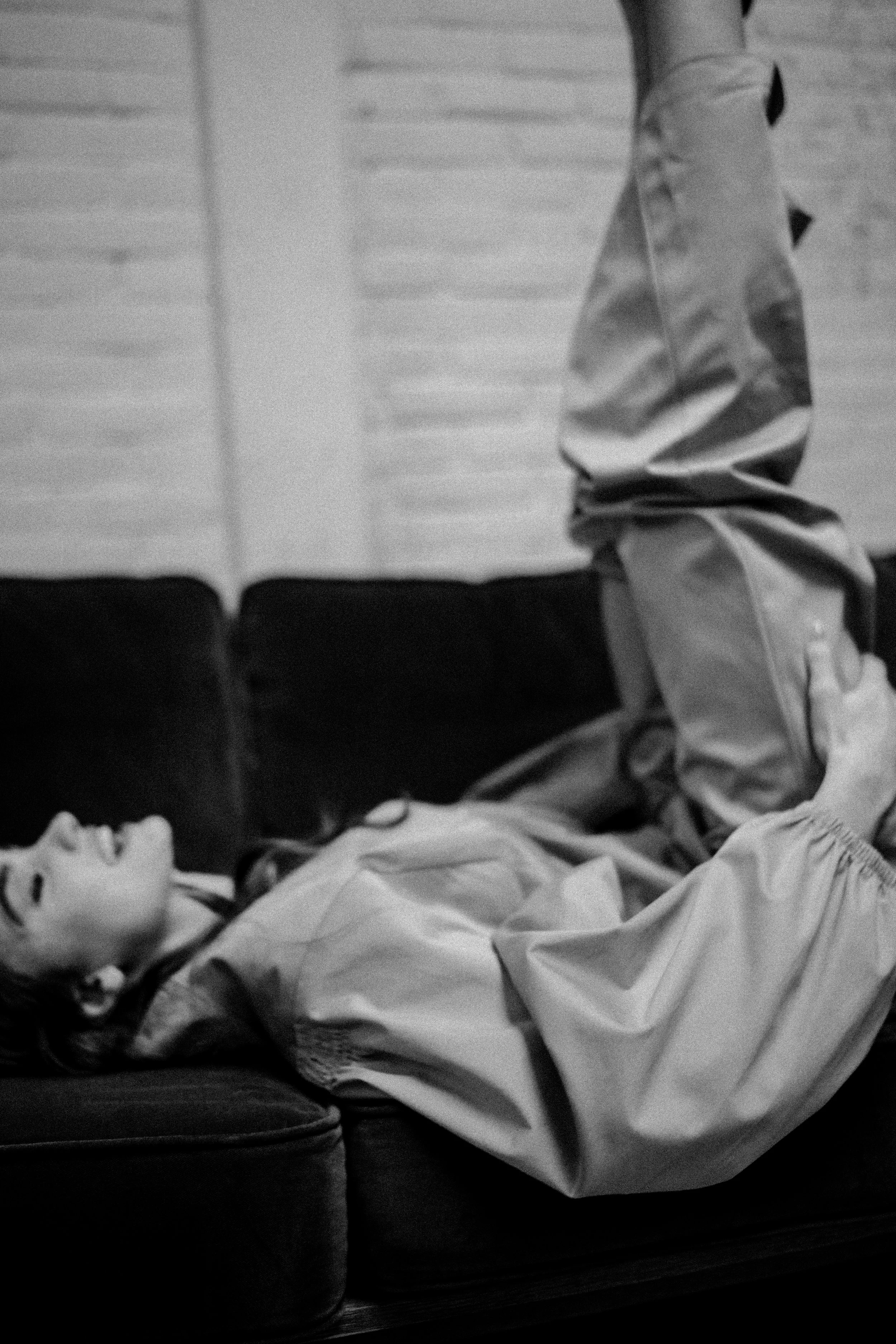 Een zwart-wit foto van een vrouw die op haar rug op een bank ligt, met haar benen omhoog en gekruist. Ze lijkt te ontspannen of te slapen, en de achtergrond heeft miniatuur raampanelen of luiken.