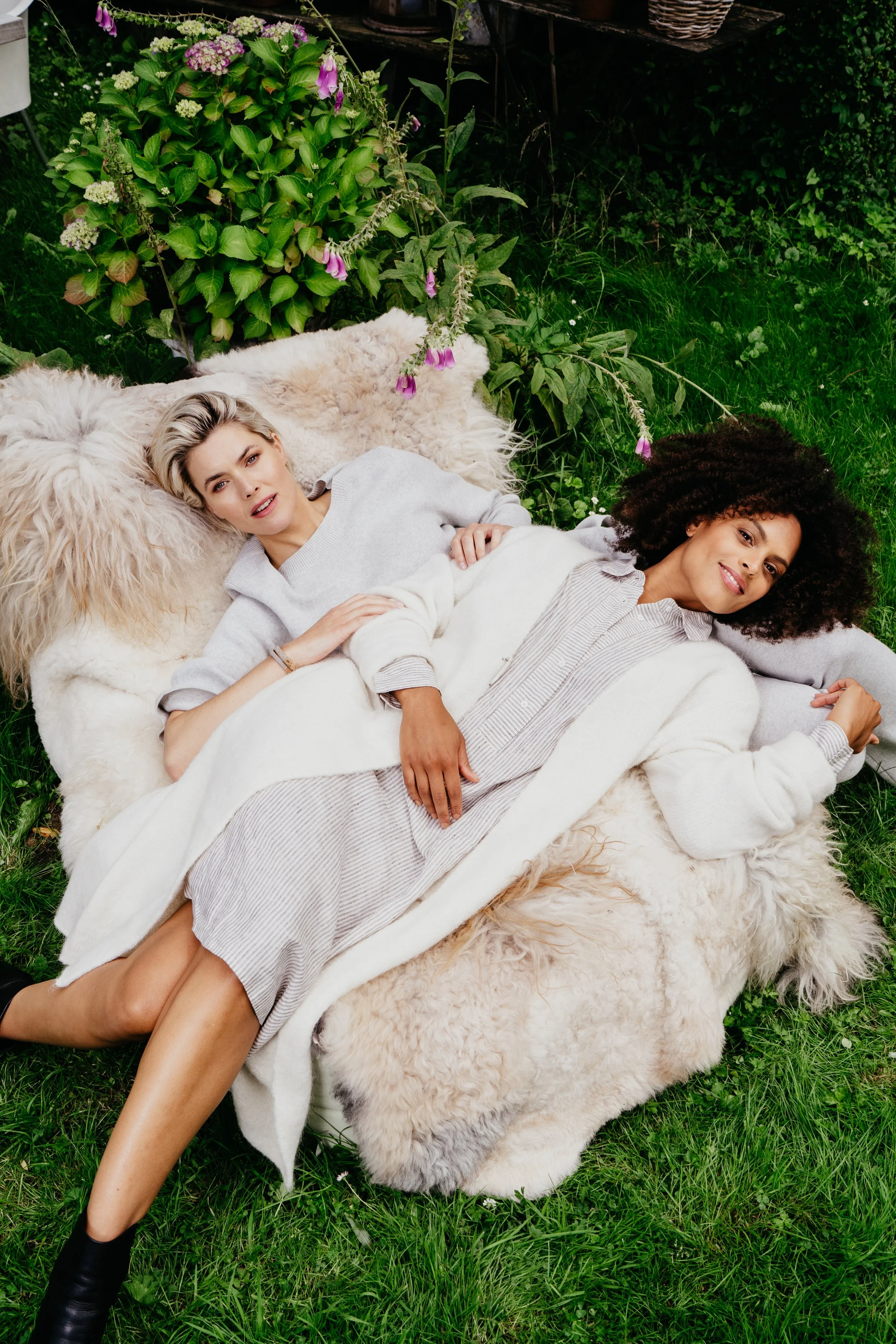 Twee vrouwen liggen op een zachte, wolkachtige deken buiten in een tuin, omgeven door groene planten en bloemen.