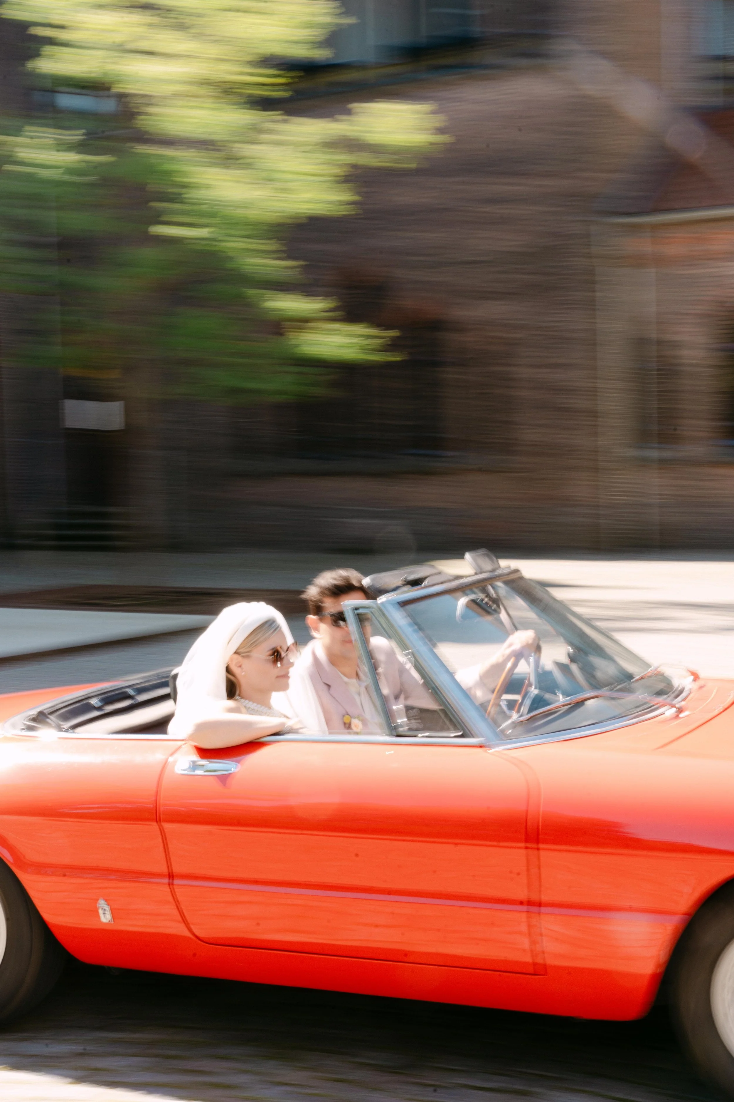 Twee mensen rijden in een rode cabriolet, de vrouw draagt een witte hoed en jurk en de man draagt een witte jas, beiden dragen zonnebrillen.