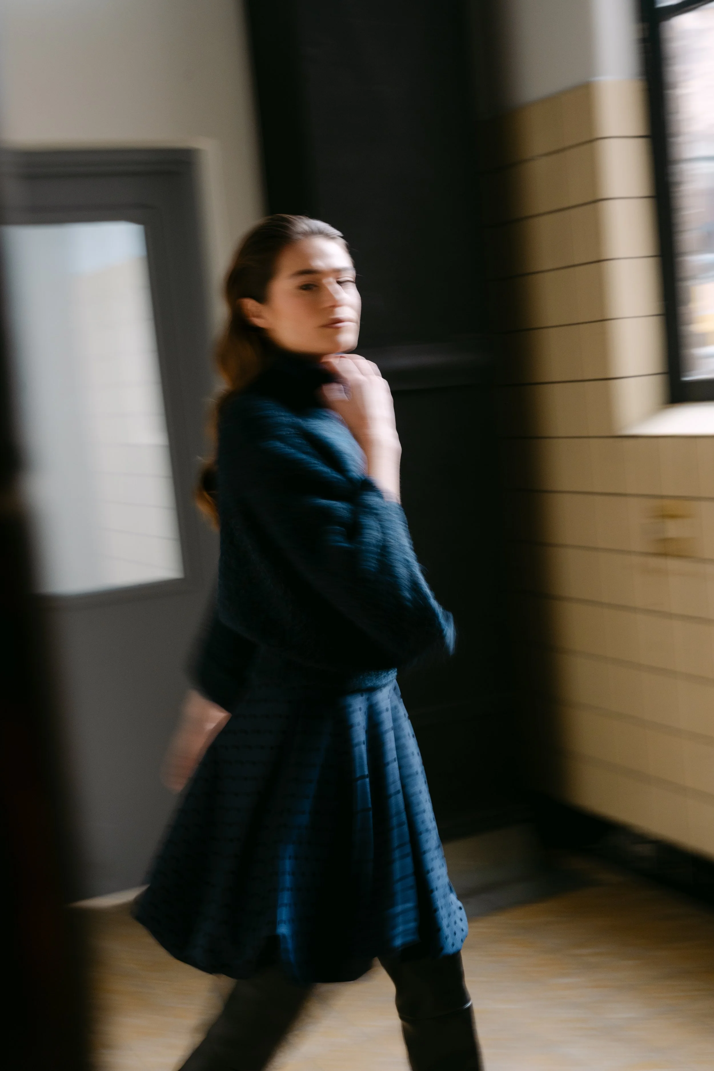 Een vrouw in een blauwe jurk en blazer loopt door een interieur met een raam en houten wanden.