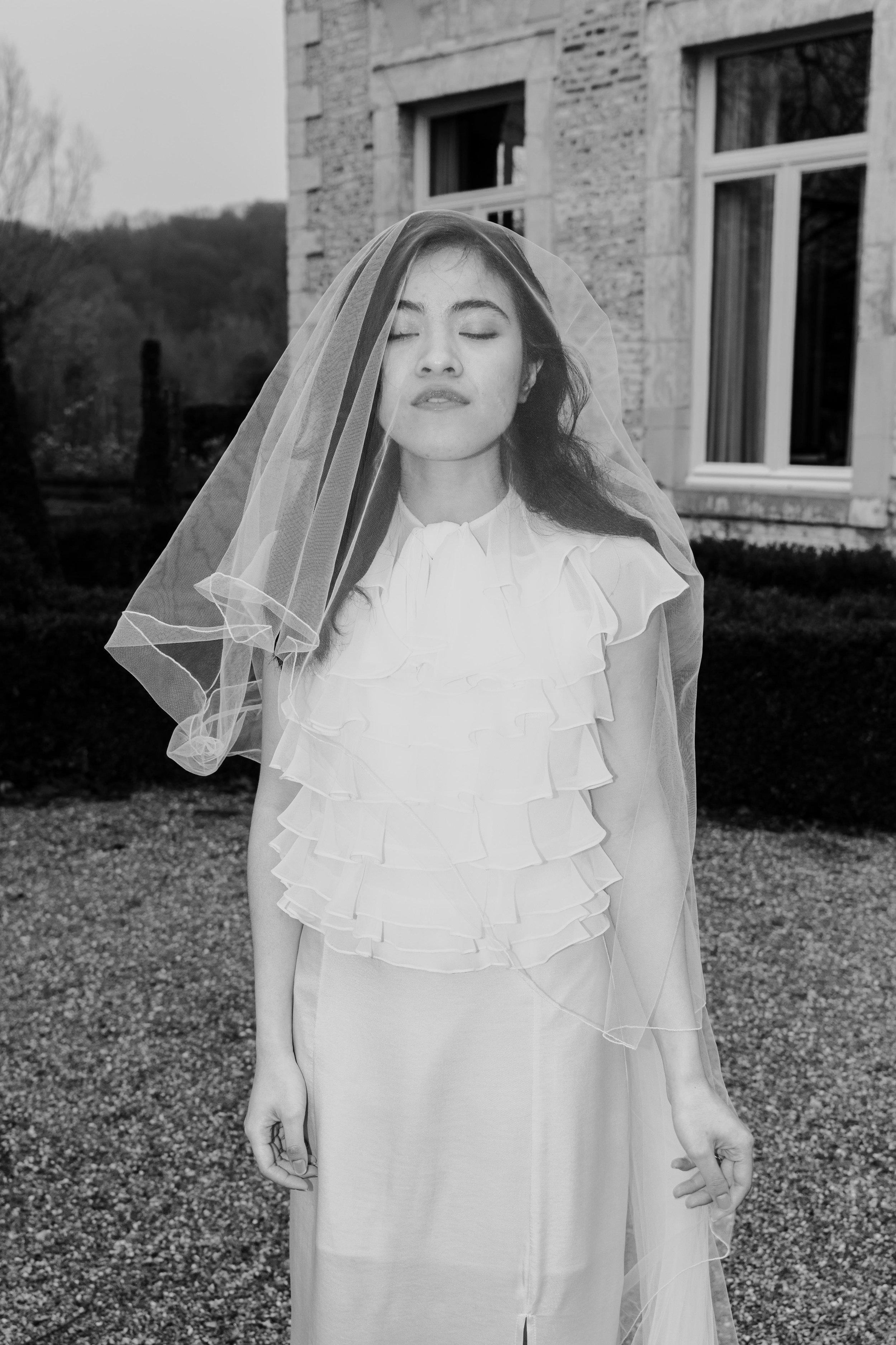 Zwarte en witte foto van een vrouw met haar ogen dicht onder een lichte sluier, staand buiten voor een stenen gebouw.
