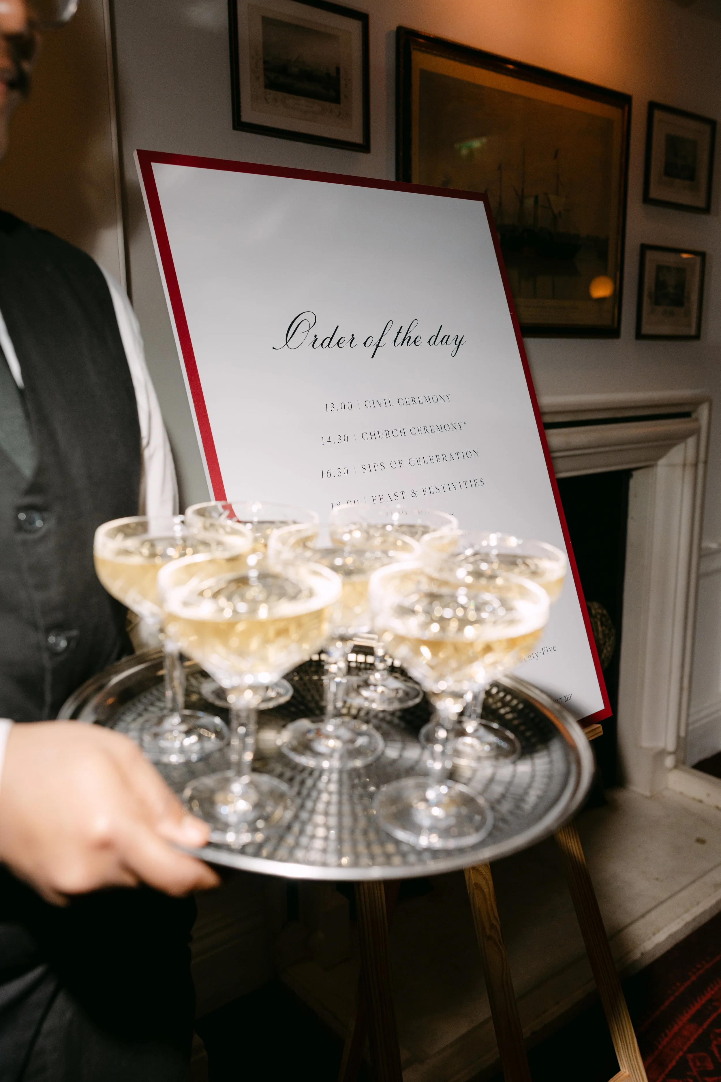 Een dienblad met glazen champagne of mousserende wijn wordt door iemand vastgehouden. Op de achtergrond is een wit bord met het schema voor de dag te zien, met een lijst van evenementen en tijden.