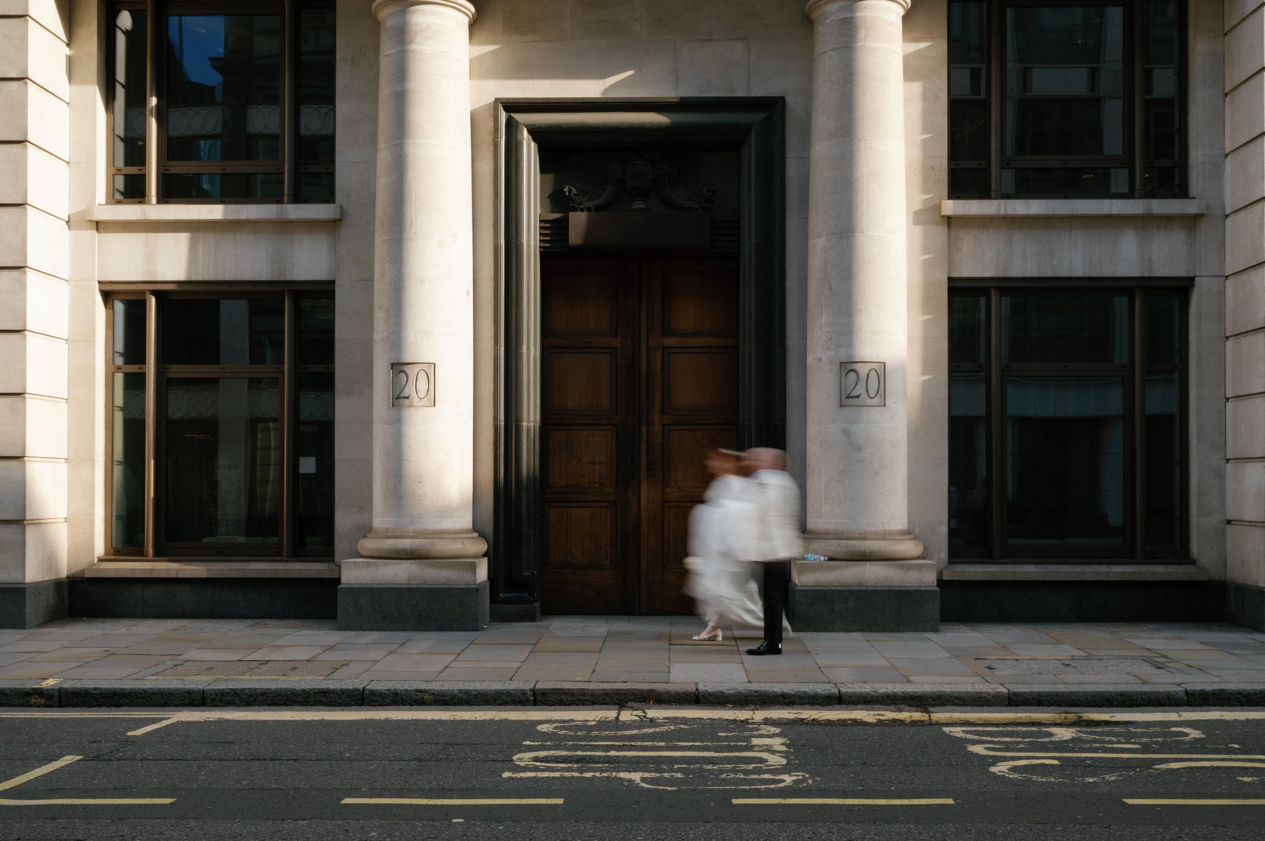 Twee mensen lopen voorbij een grote houten deur in een gebouw met klassieke architectuur en witte zuilen, nummer 20 op de gevel.