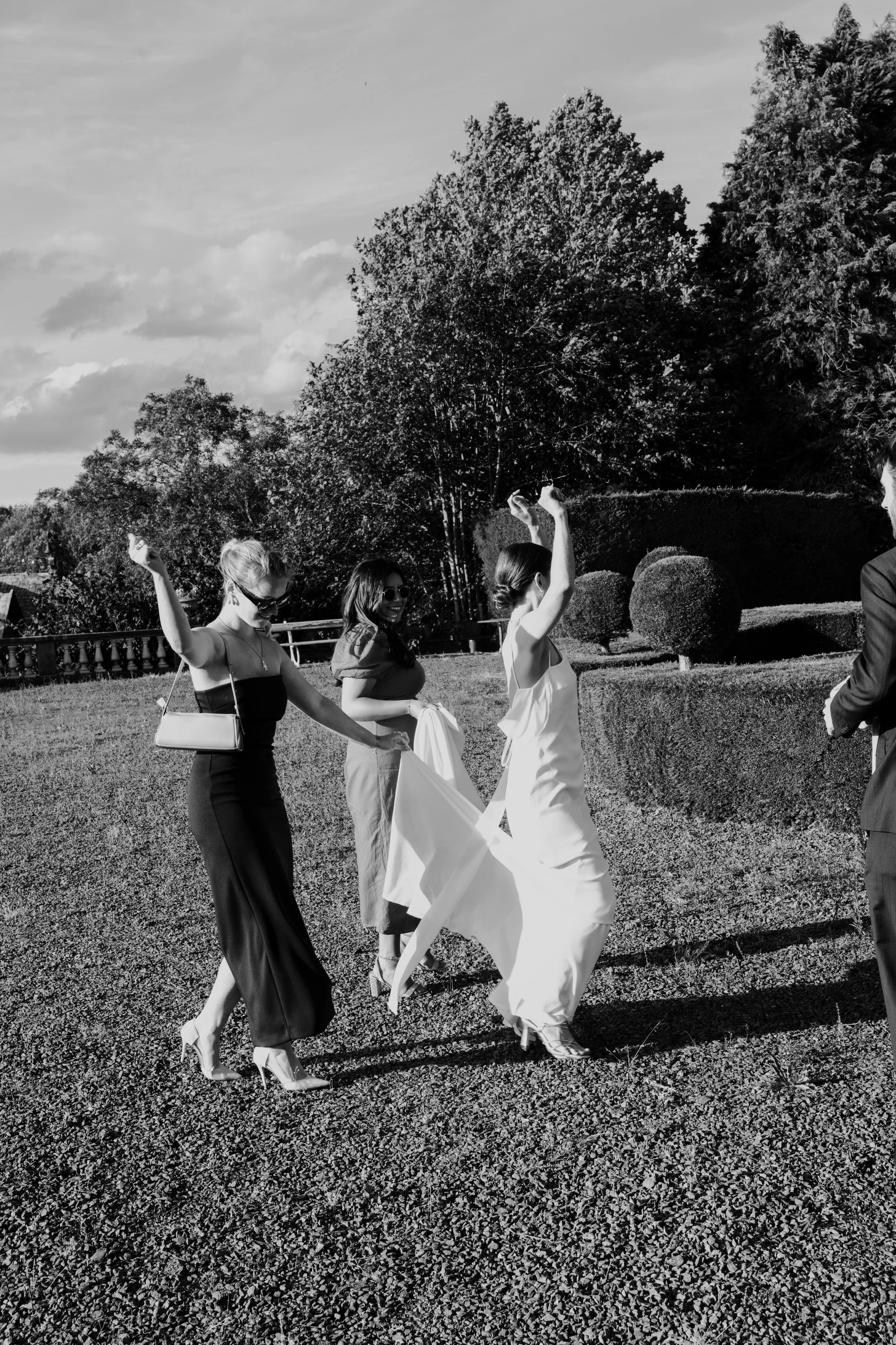 Vrouwen in elegante jurken die zich voorbereiden voor een bruiloft buiten, met een tuin en bomen op de achtergrond.