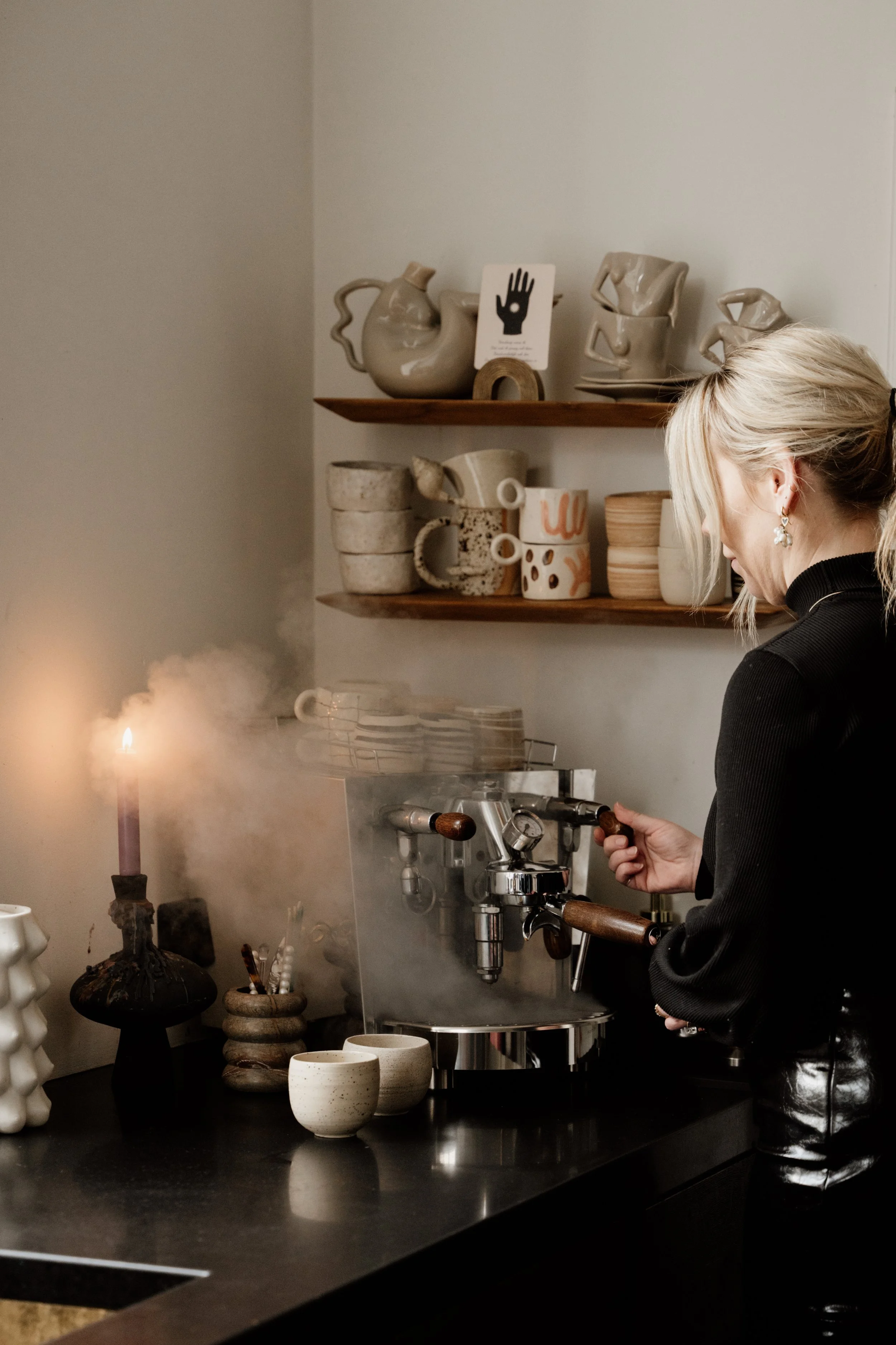 Een vrouw bereidt koffie met een espressomachine in een keuken, met keramische kopjes en vazen op een zwarte keukentafel en houten planken met keramiek op de achtergrond.