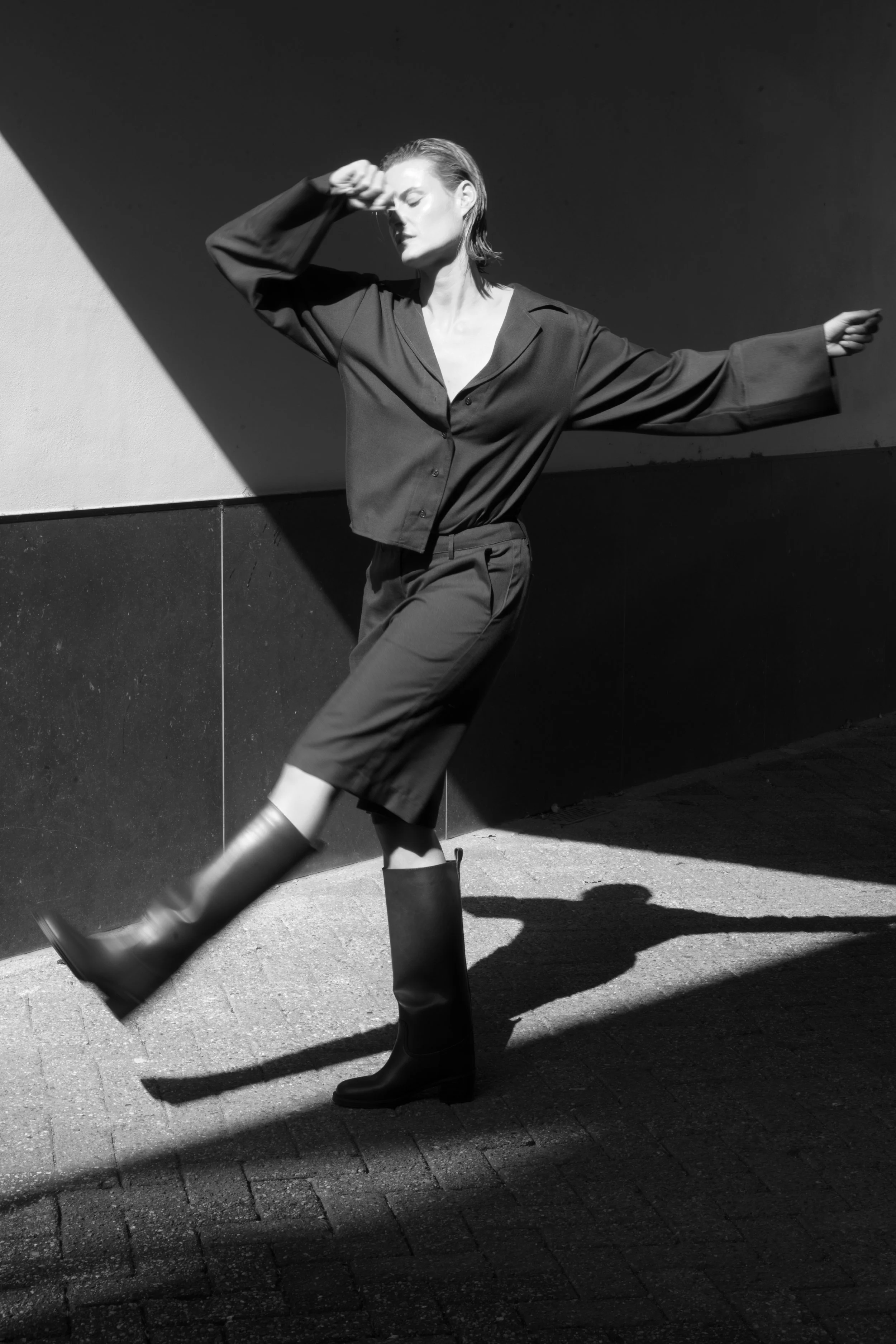 Zwart-wit foto van een vrouw die buiten staat, gekleed in een losse blouse, culottes en regenlaarzen. Ze houdt haar hand voor haar gezicht en lijkt de zon te vermijden.