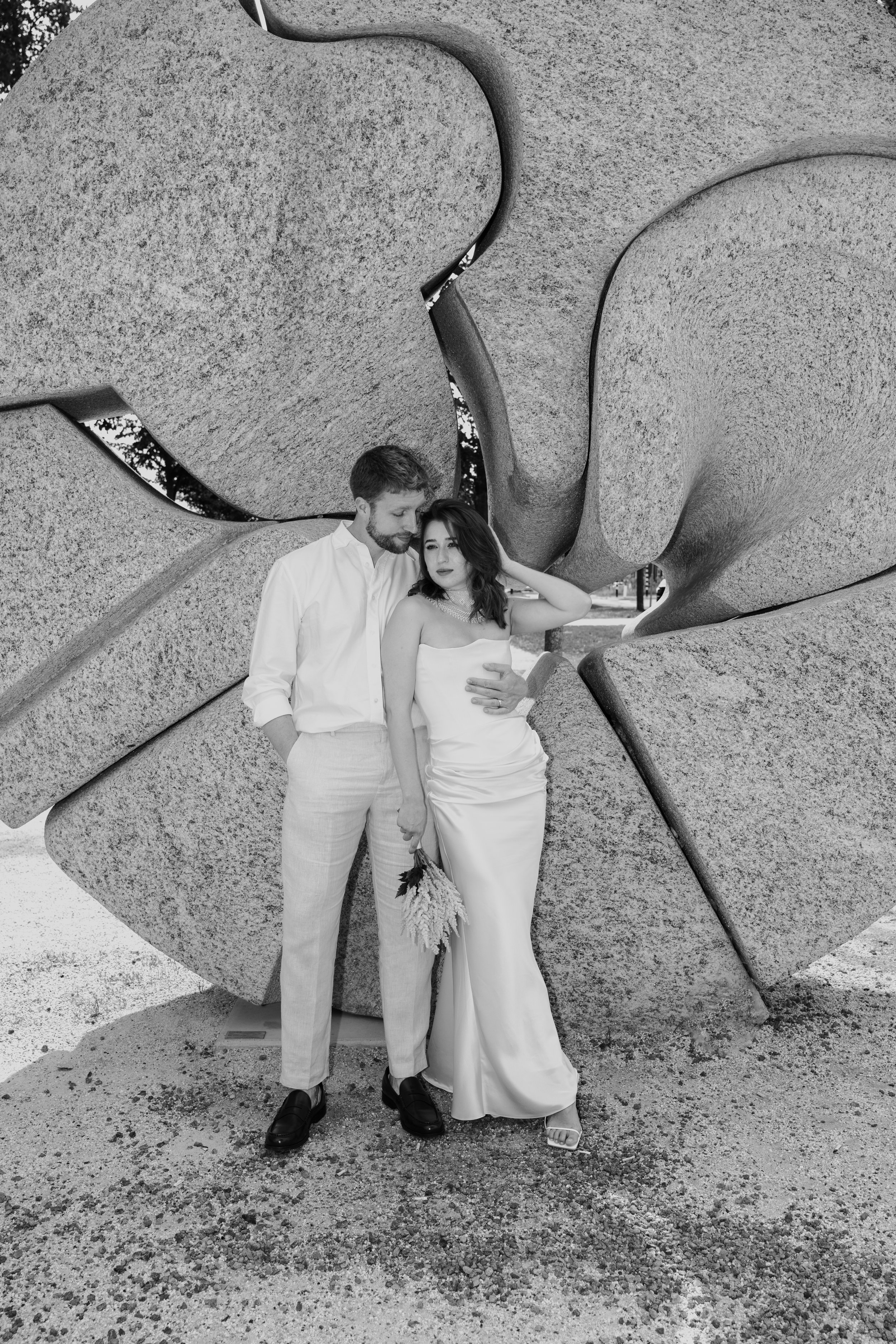 Een zwart-wit foto van een man en vrouw in formele kleding die dicht bij elkaar staan voor een grote abstracte betonnen sculptuur.