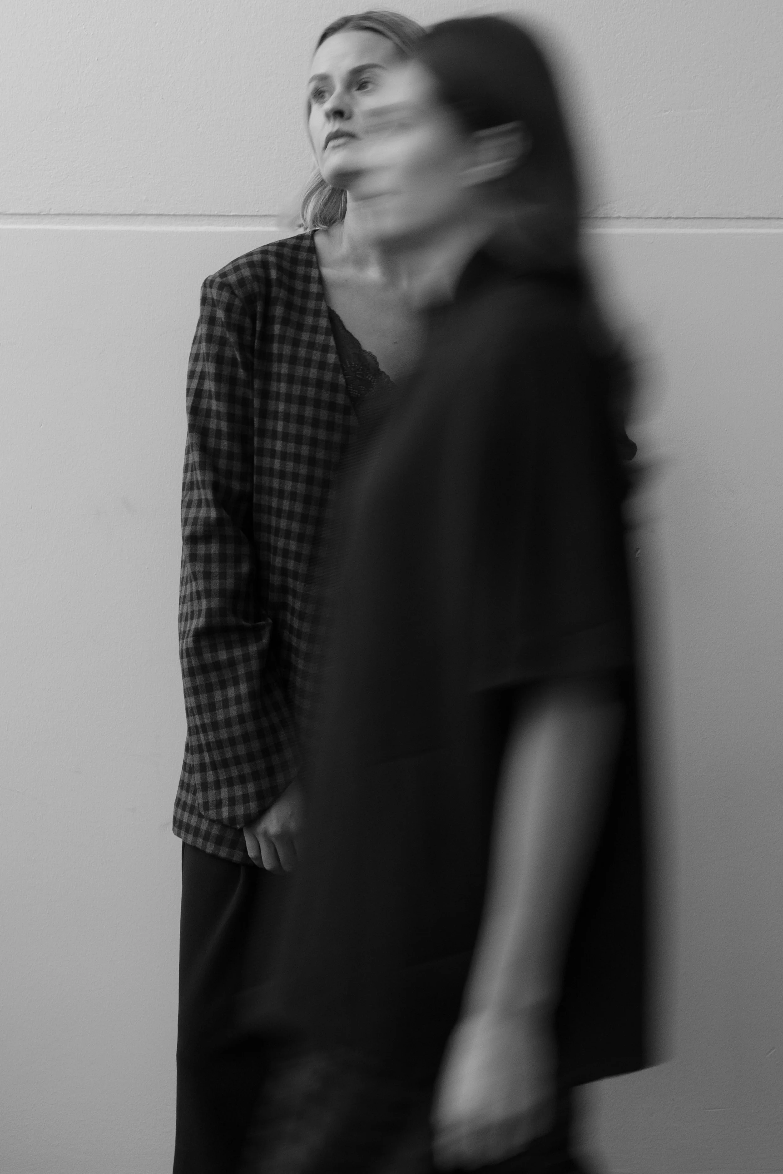 Twee vrouwen staan tegen een lichtgekleurde muur, één met lang haar en een geruite jas, de andere met kort haar en een donkere jas, beide in zwart-wit en de vrouw met kort haar lijkt in beweging.