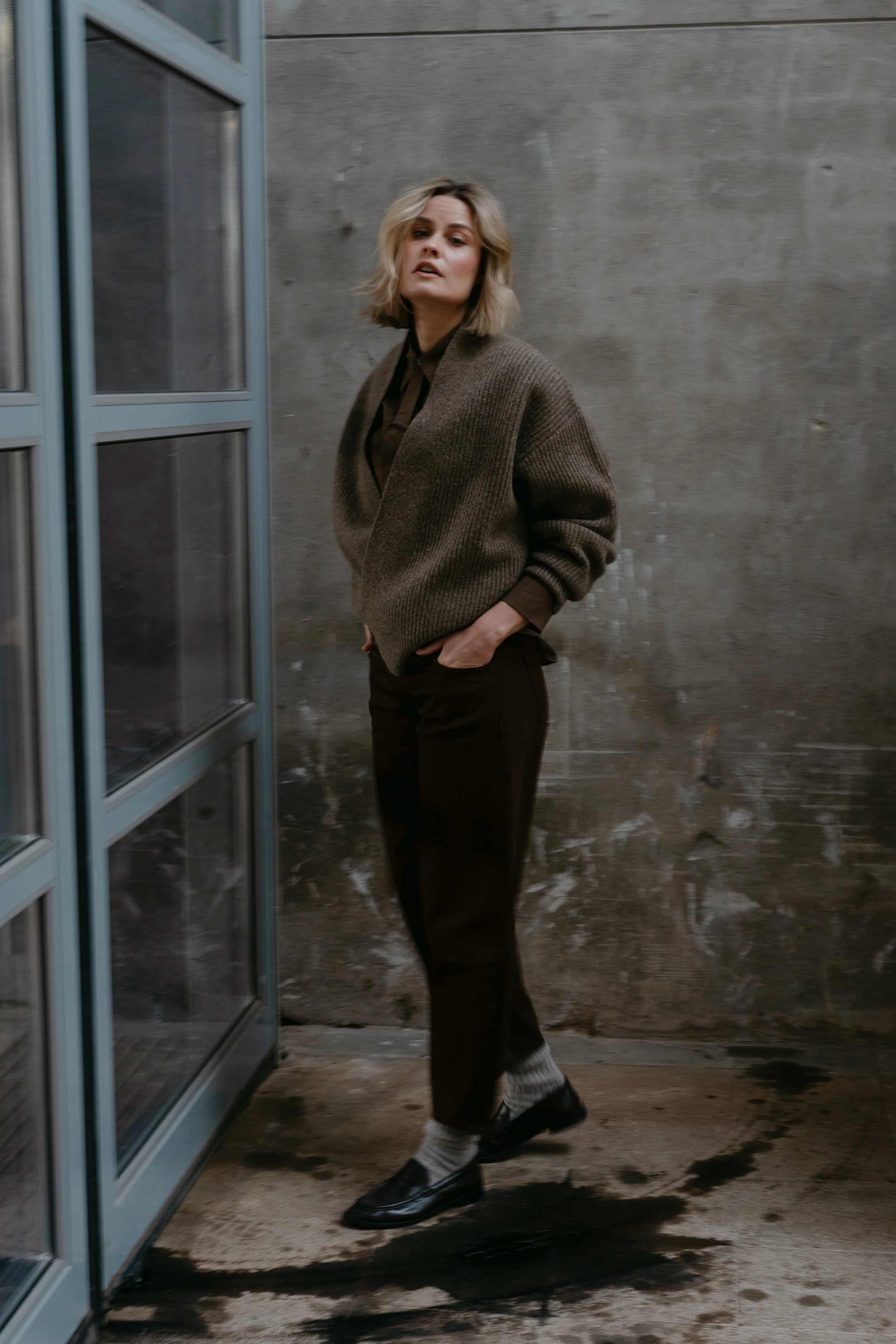 Vrouw met blond haar draagt een bruine trui en zwarte broek, staat binnen bij een glazen deur, met een stenen muur achtergrond.