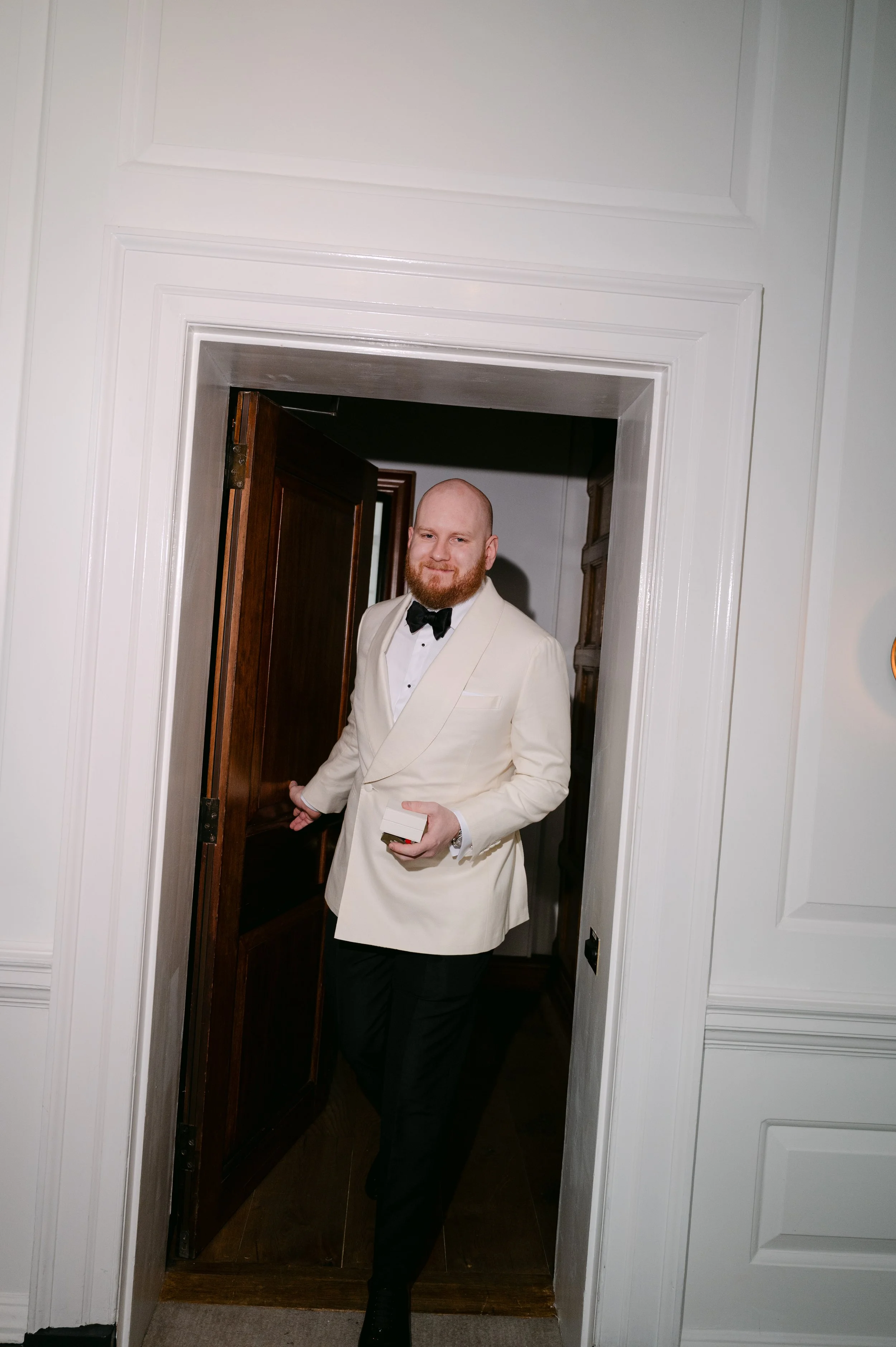Man in wit tuxedo en zwarte strik laat door de deur lopen, houdt een small box in zijn hand.