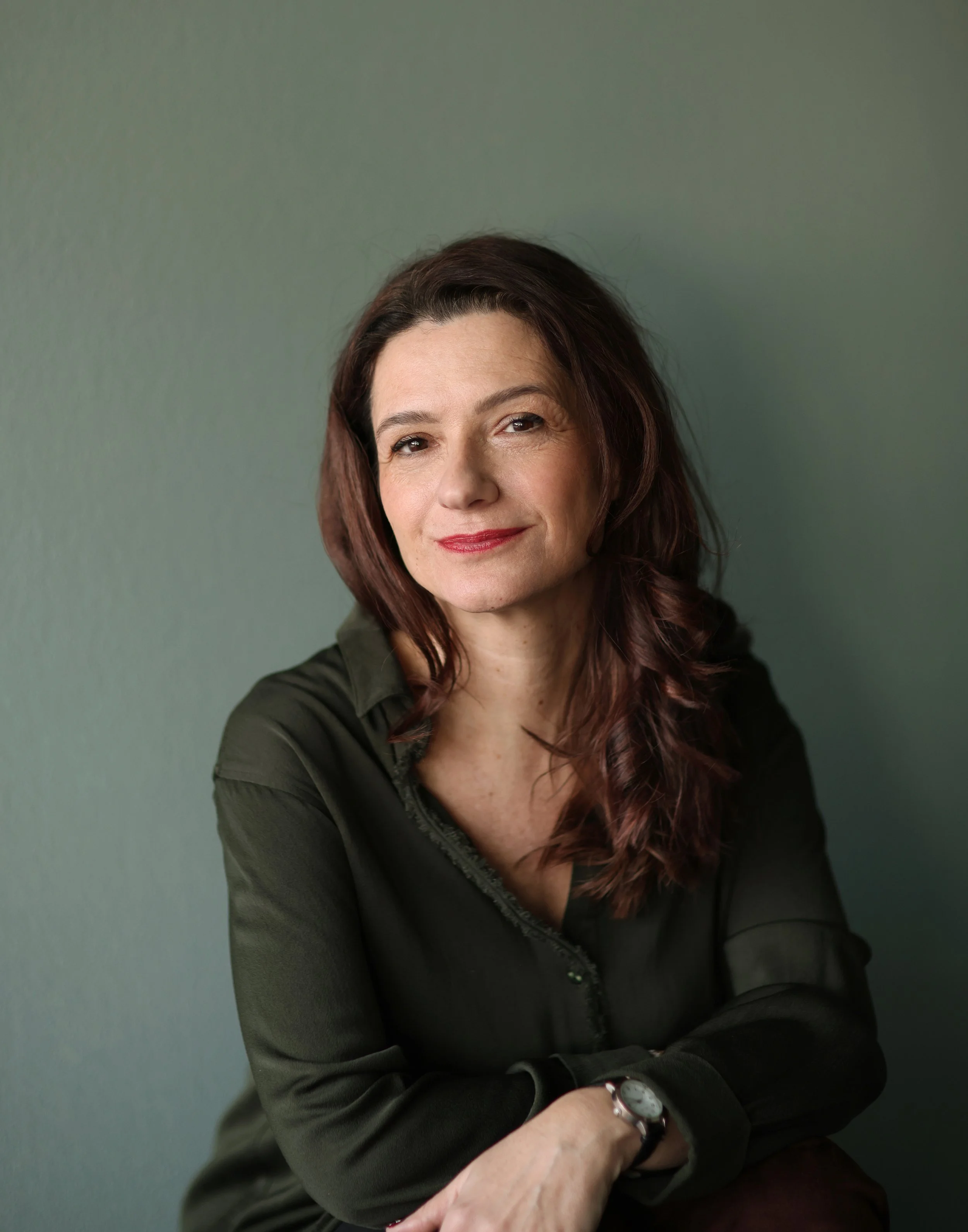 Portrait d'une femme souriante aux cheveux bruns, portant une chemise noire, assise contre un mur vert.