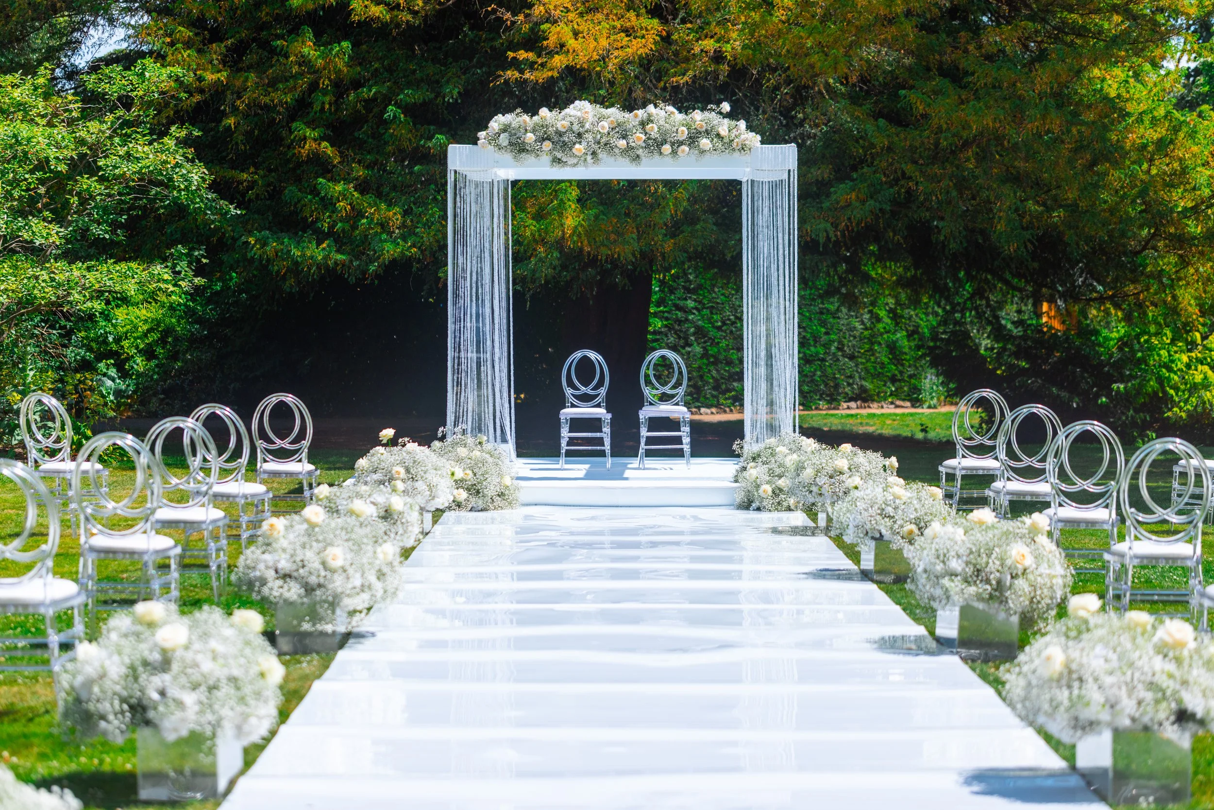 Outdoor summer wedding ceremony with white floral arch and aisle arrangements in a garden