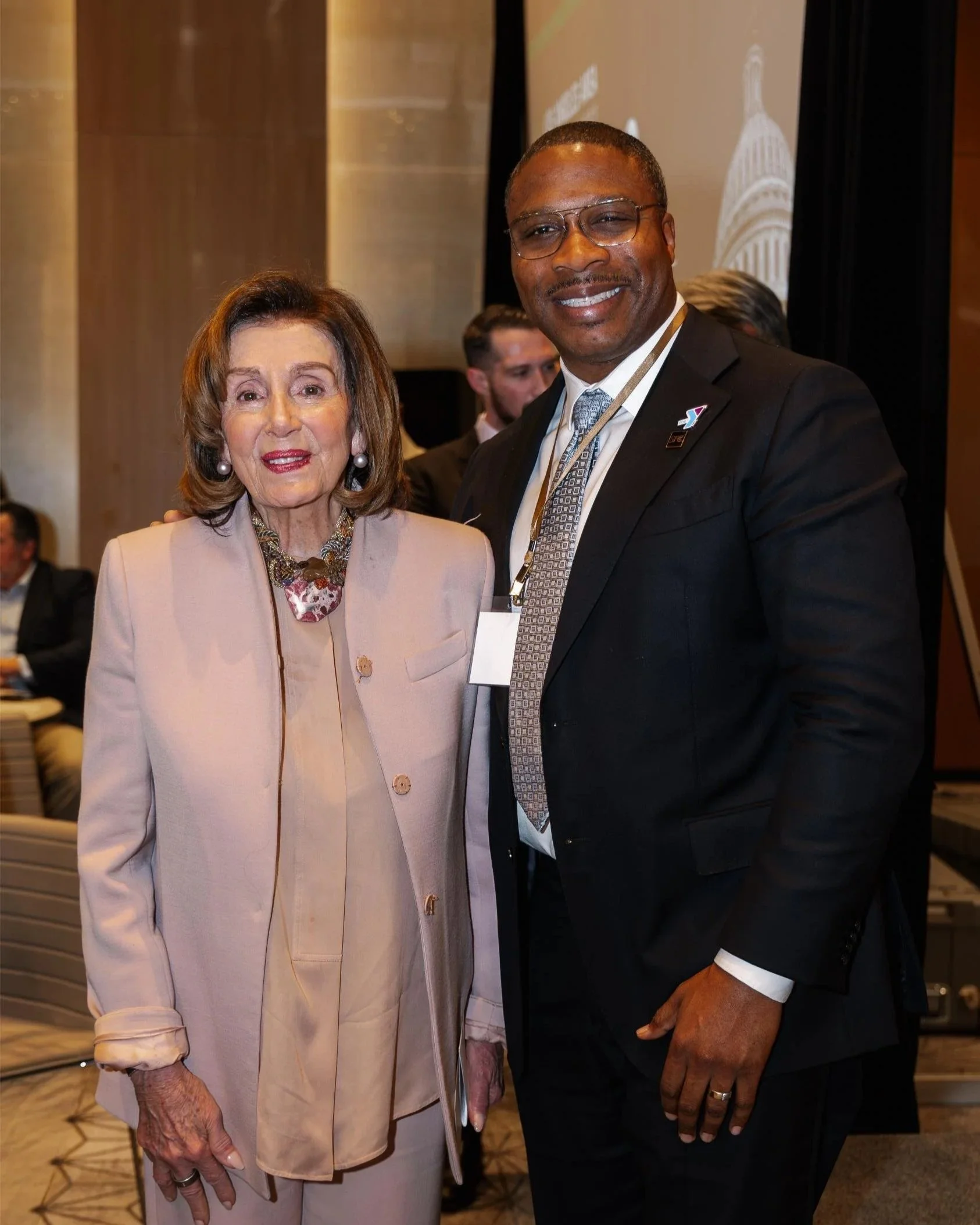 A woman and a man in business attire posing together at an event, with a presentation screen and other people in the background.