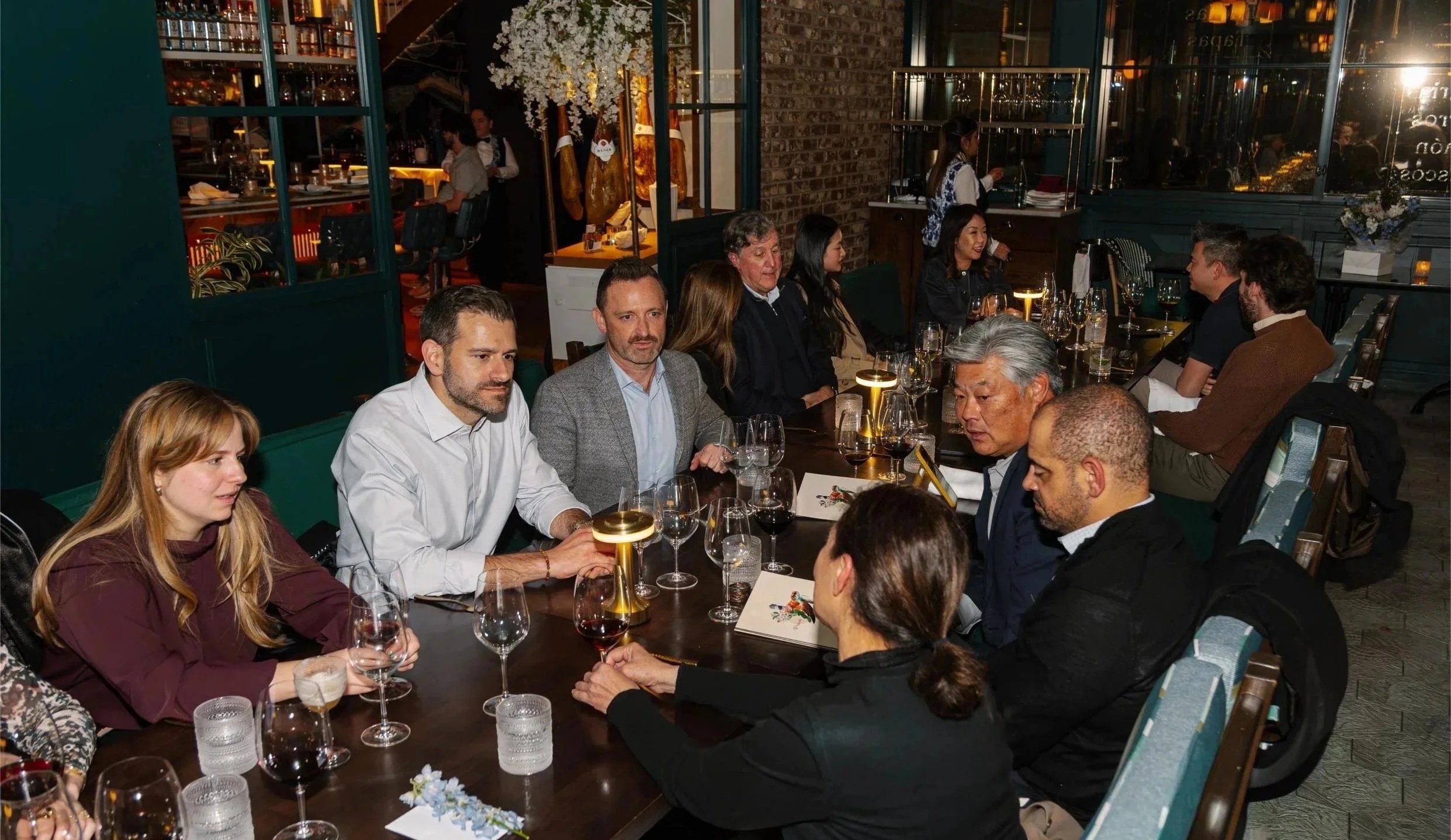 A group of people sitting around a dinner table in a restaurant, engaged in conversation with wine glasses and menus on the table, dim lighting, and a decorated background.
