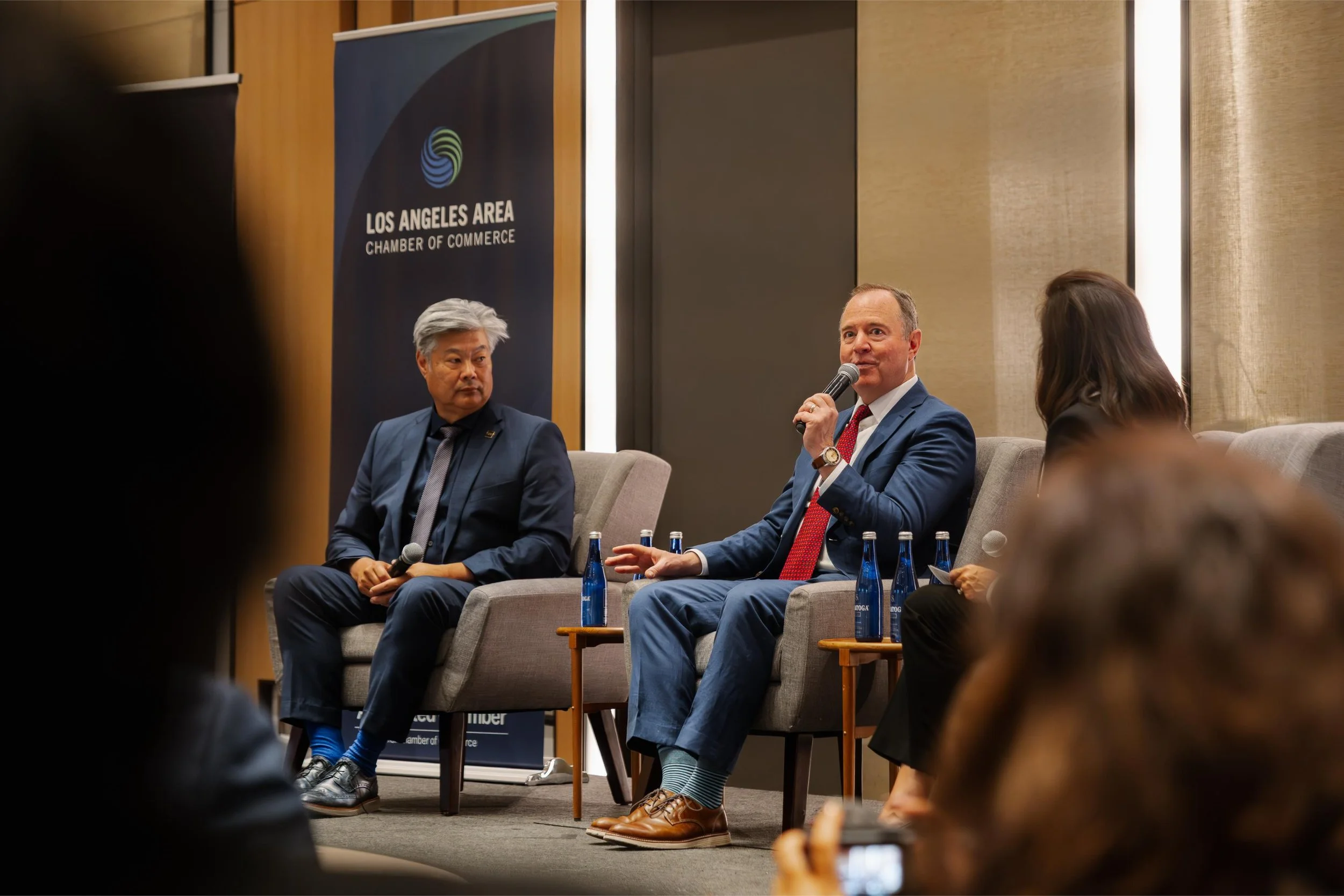 A panel discussion at the Los Angeles Area Chamber of Commerce with three people seated on stage, including a man speaking into a microphone, and an audience in front.