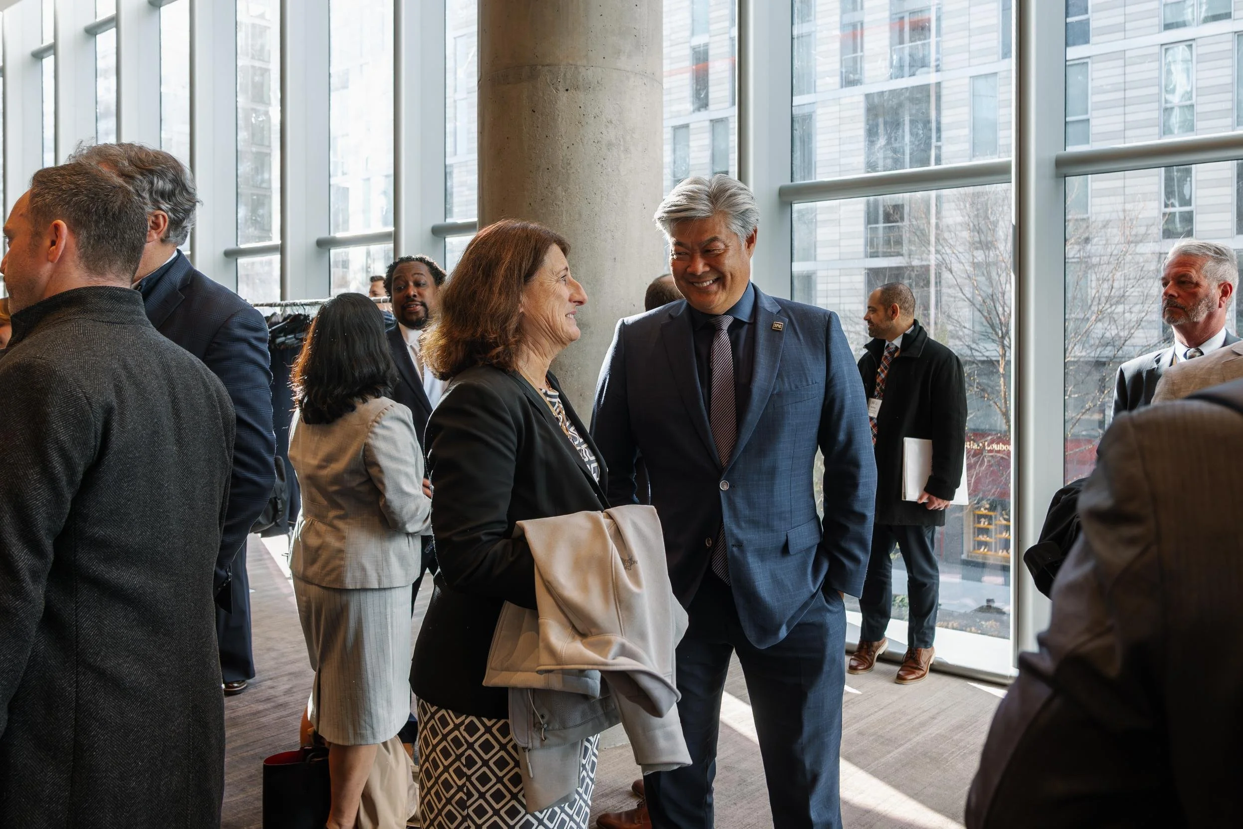 Two people, a woman and a man, are smiling and talking at a conference or business event. The woman is holding a beige coat, and the man is wearing a blue suit. Other attendees are visible in the background near large windows in a modern building.