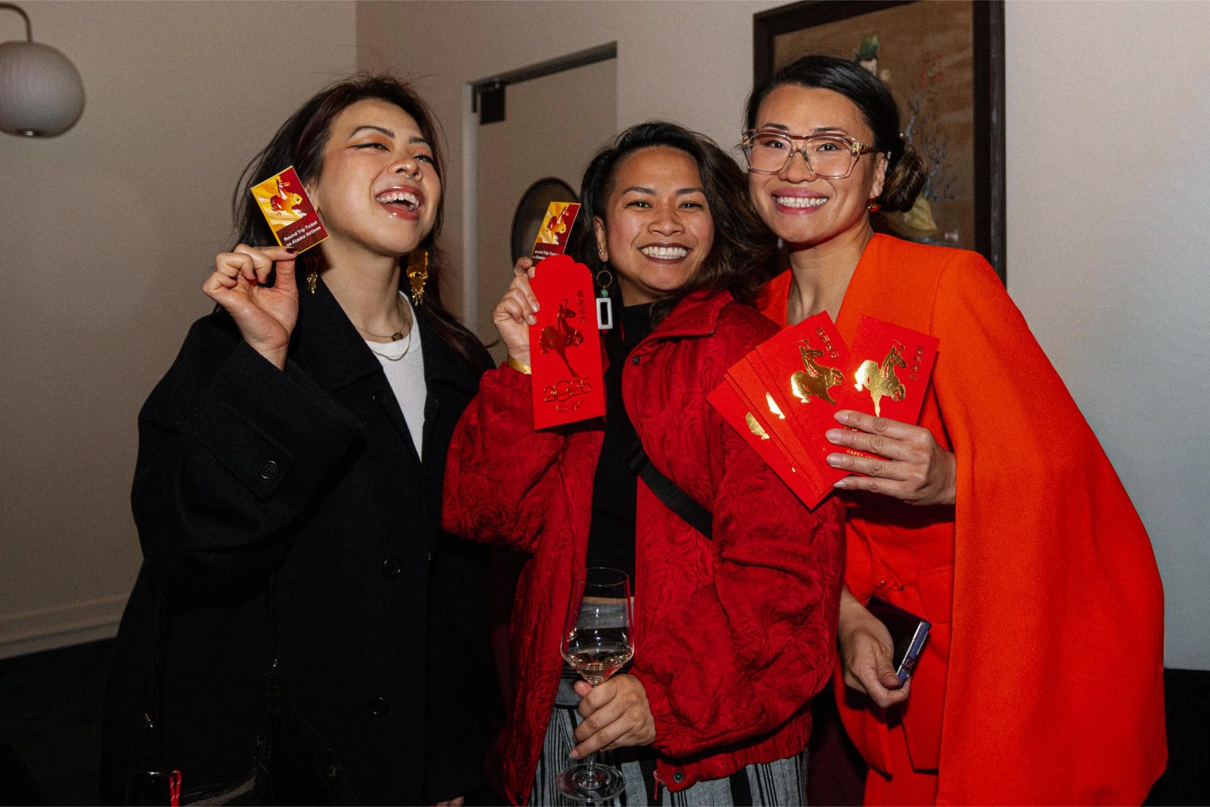 Three women celebrating Chinese New Year indoors, holding red envelopes and a glass of wine, dressed in festive red and black attire.