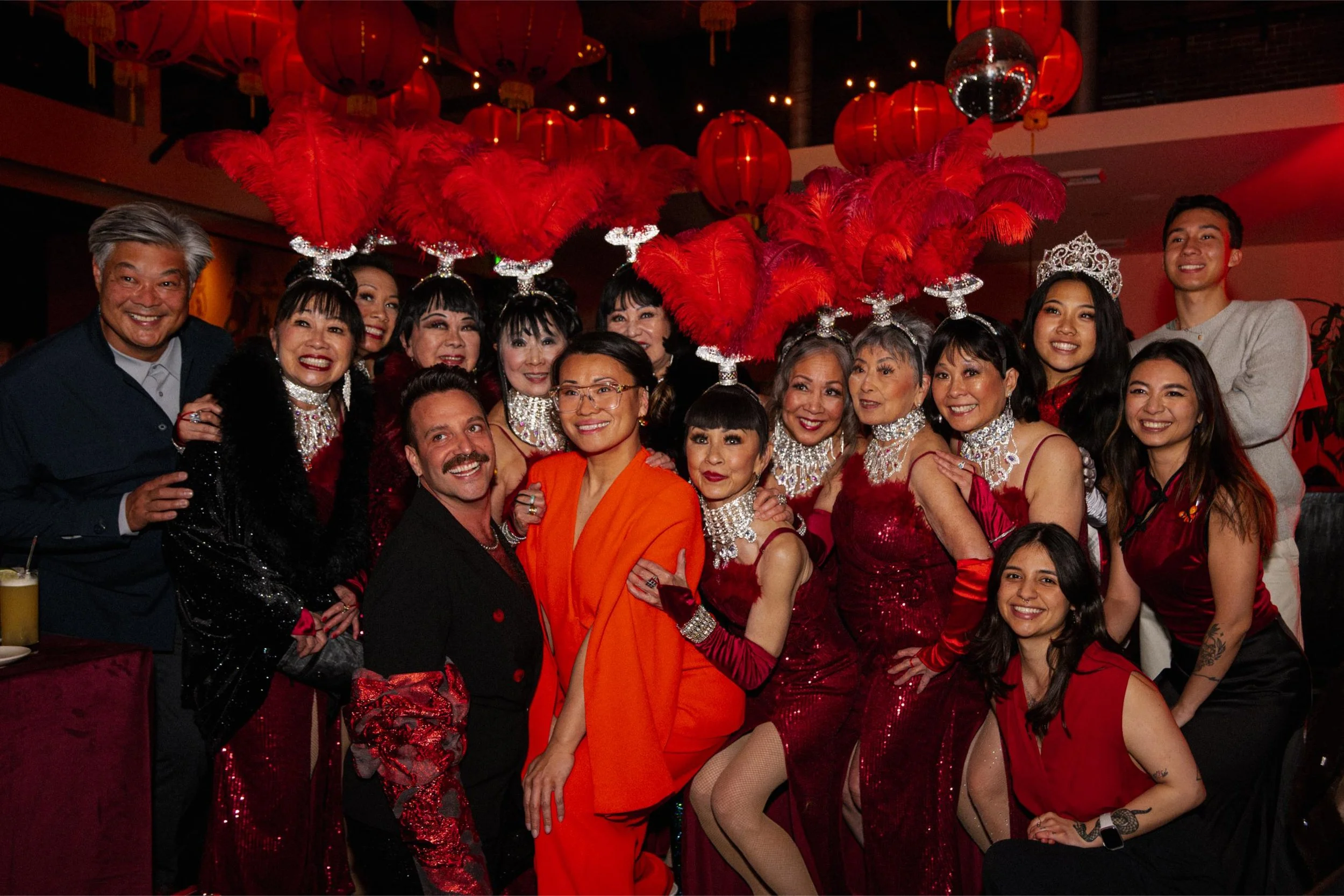 A group of people at a celebration wearing red and black clothing, some with feathered headpieces and jewelry, in a festive setting with red lanterns and a disco ball hanging from the ceiling.
