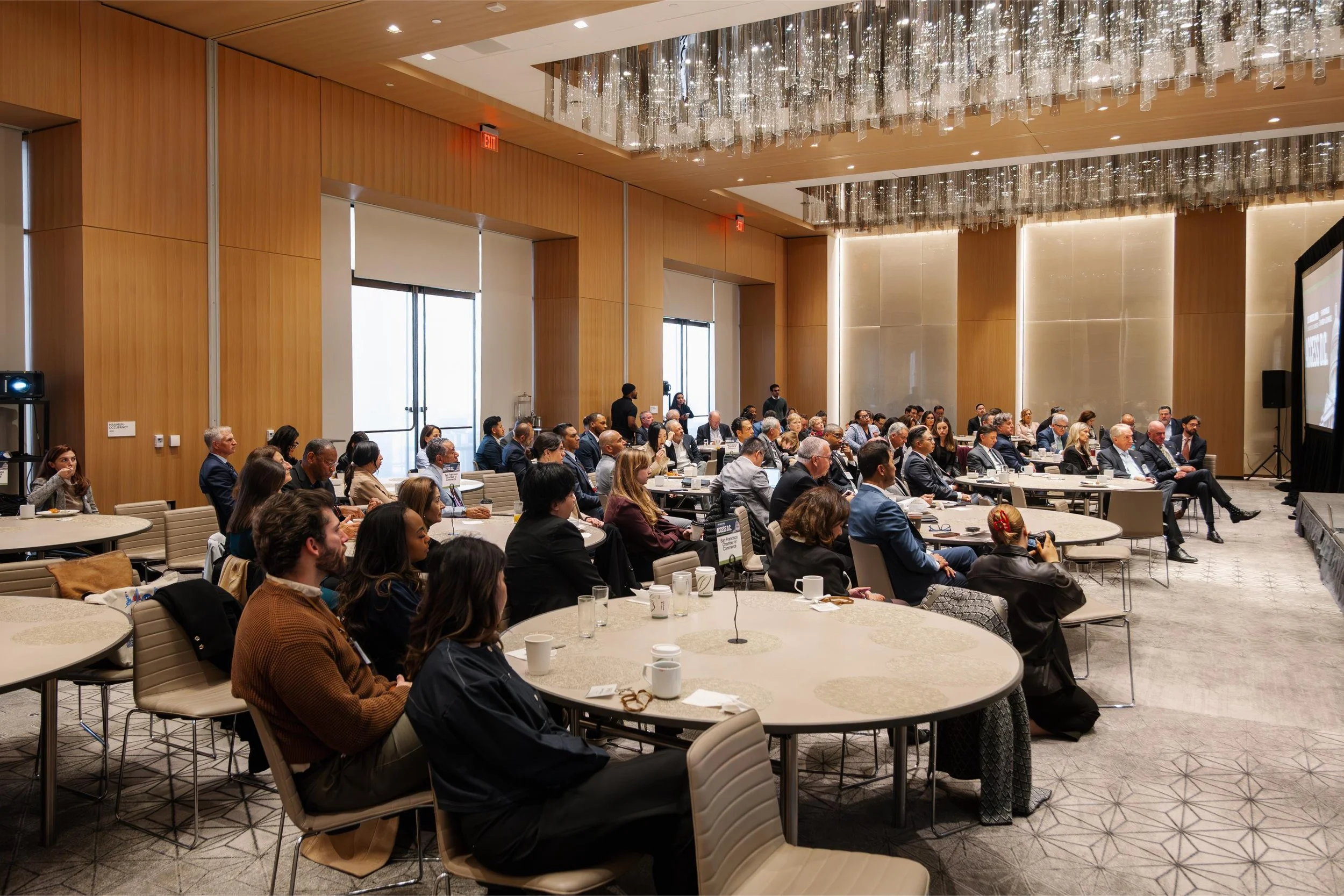 An audience seated at round tables in a conference room, attentively watching a presentation on a large screen to the right.