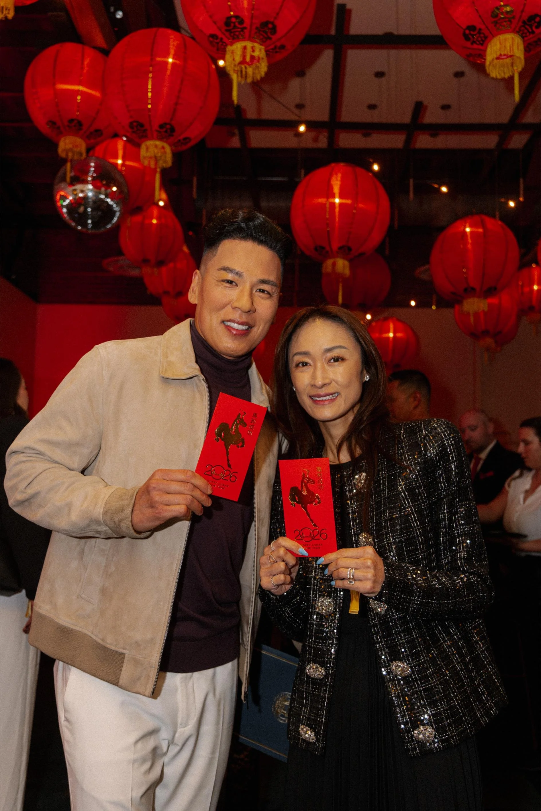 Two people holding red envelopes with a decorative horse and the year 2026, celebrating a Chinese New Year event under red lanterns.