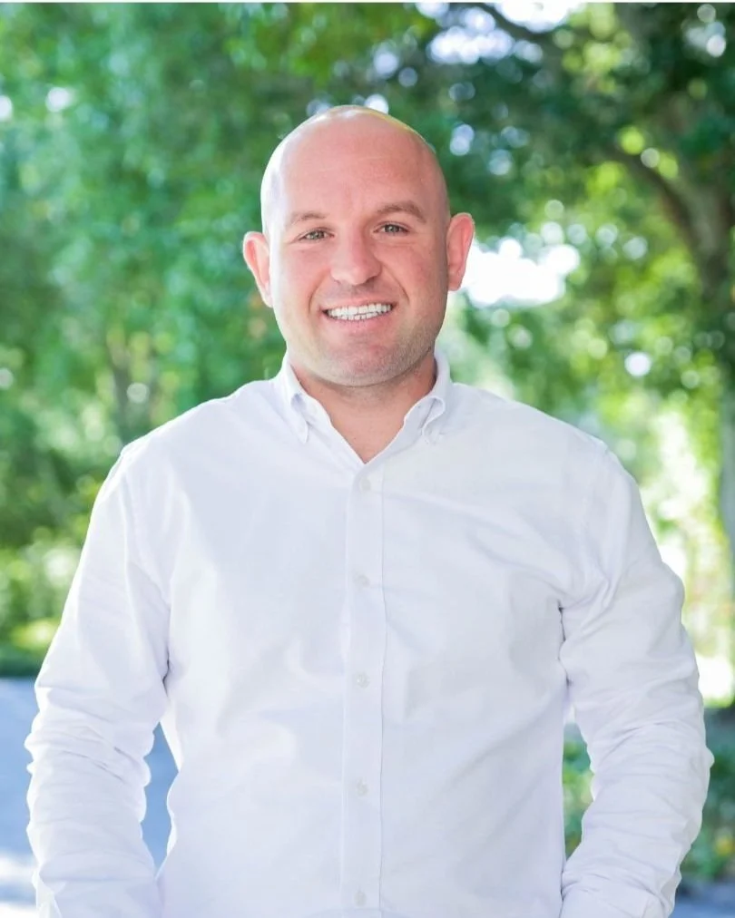 A person wearing a white button-up shirt, standing outdoors with a backdrop of green trees.