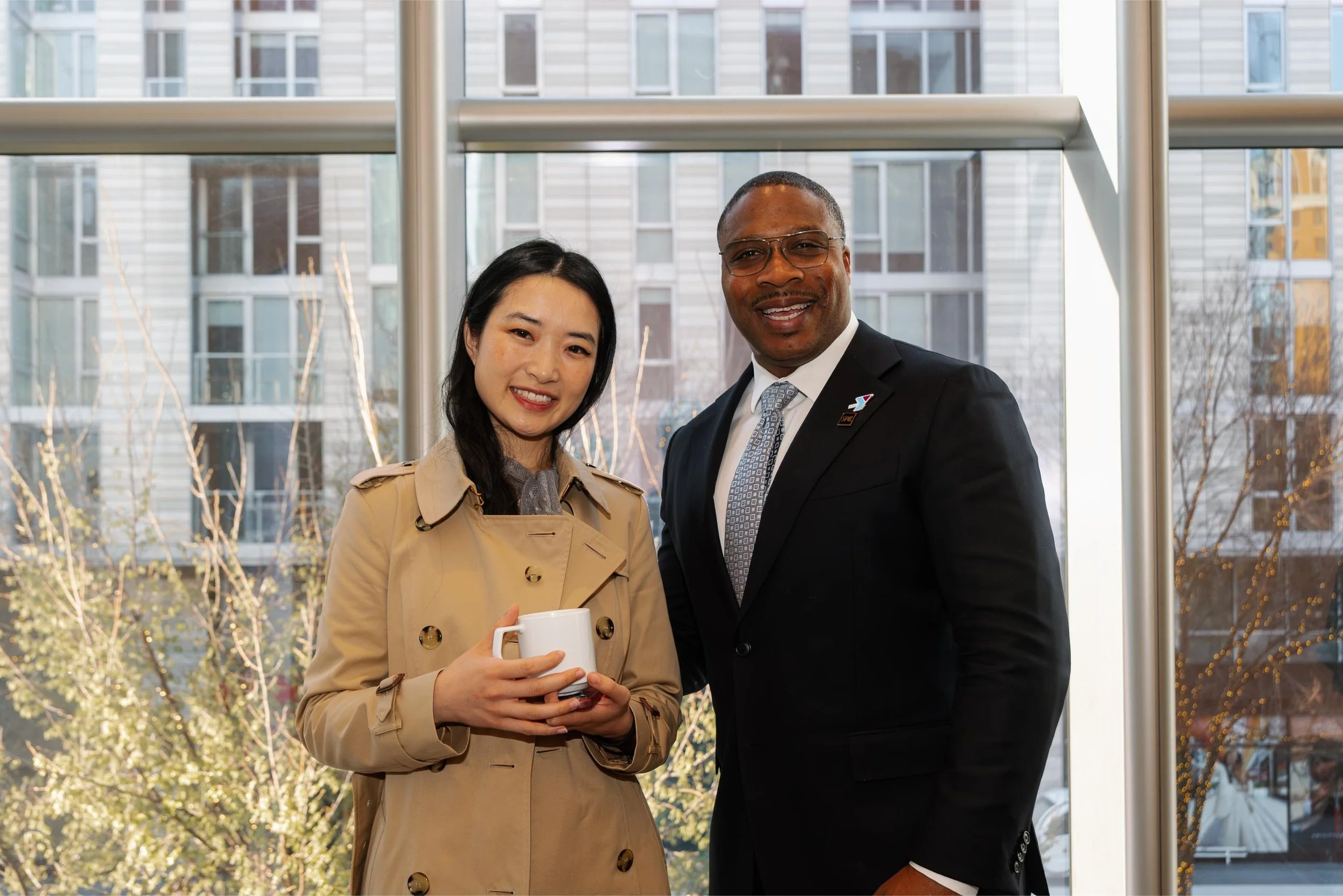 A woman and a man posing indoors in front of a large window, with outdoor buildings and trees visible through the window. The woman is holding a white coffee mug and wearing a beige trench coat. The man is dressed in a dark suit with a tie and glasse