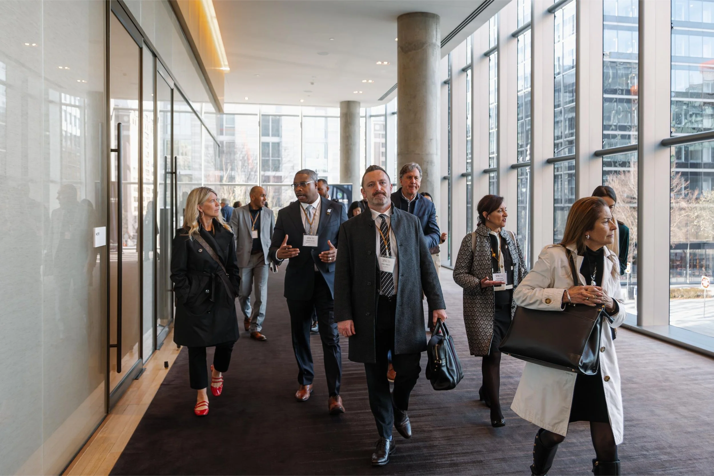 Business professionals walking through a modern office building corridor with large glass windows and cityscape view.